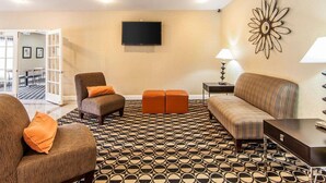 Un salon comprenant un canapé, un fauteuil, un pouf, un revêtement de sol à motifs, un téléviseur, une lampe et un élément décoratif mural.