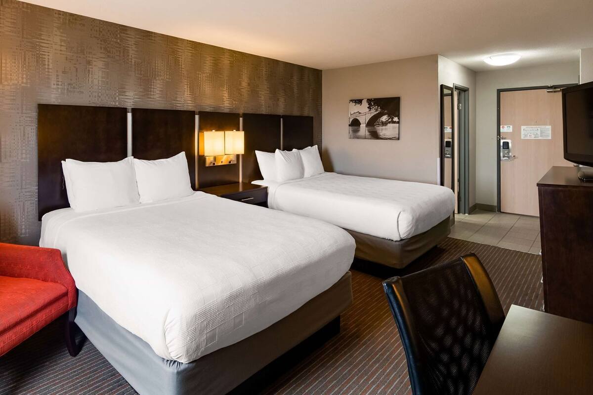 A hotel room with two beds, a red armchair, a desk, and a painting on the wall.