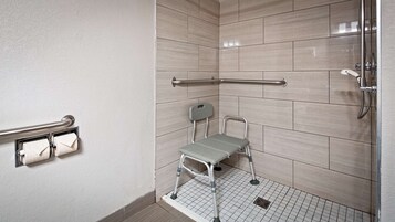 Zona de ducha con barra de apoyo, taburete y dispensador de papel higiénico.
