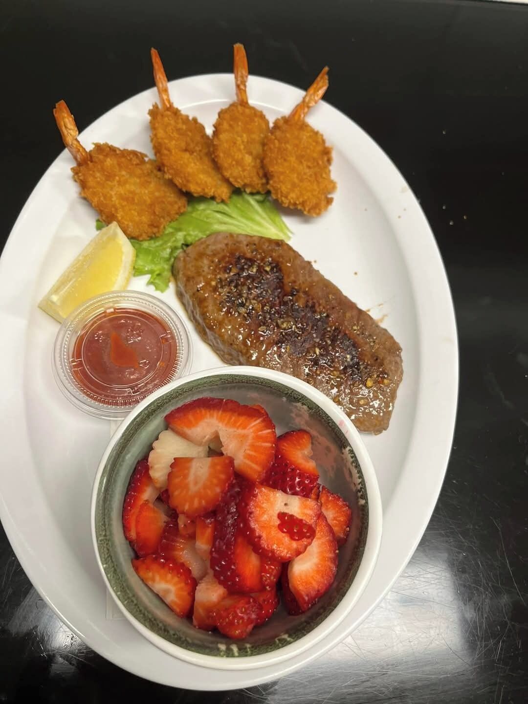 Une assiette contenant des crevettes panées, une galette de viande grillée, une salade d’accompagnement, un quartier de citron et un bol de fraises coupées en tranches.