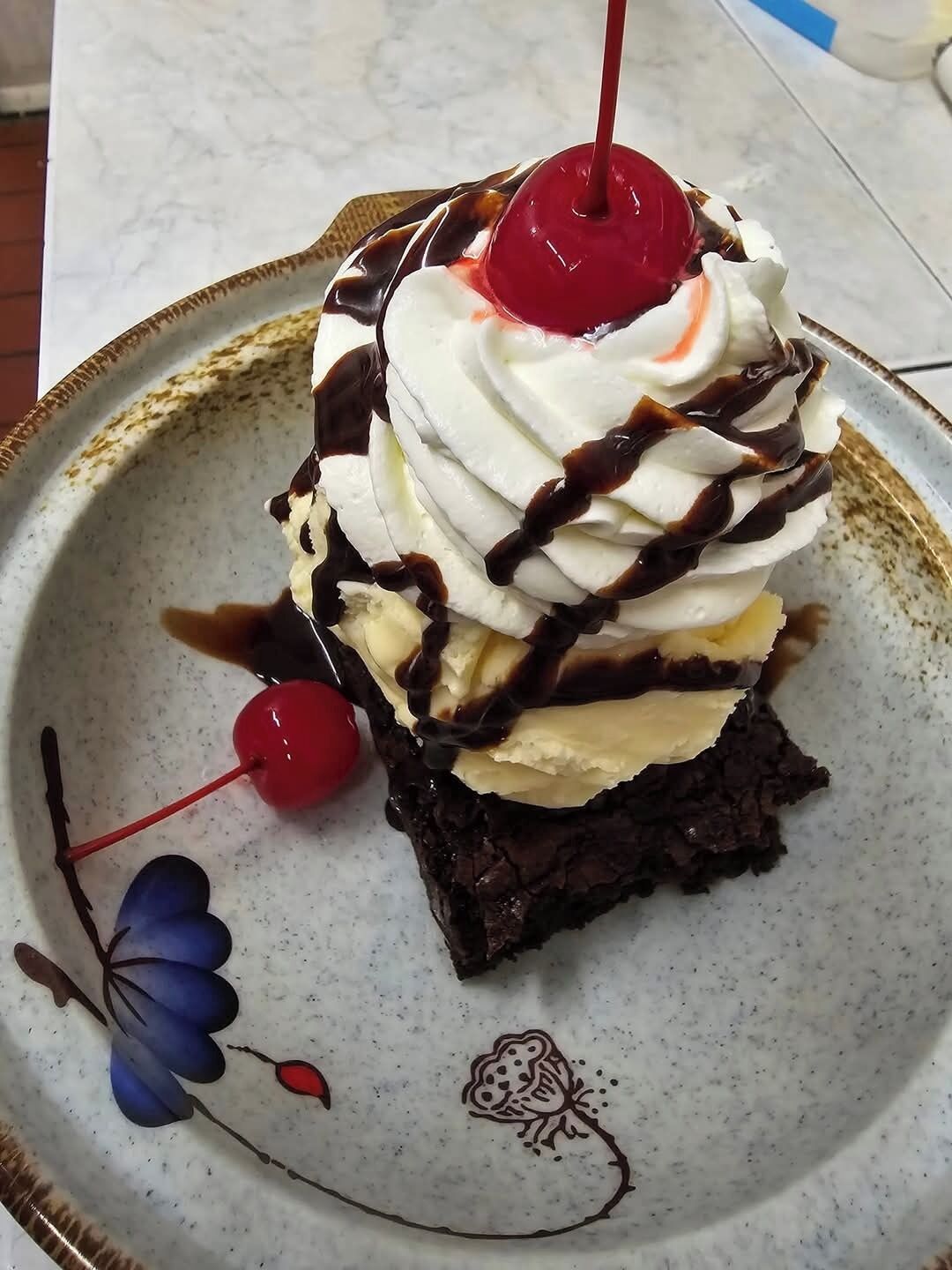 A slice of layered dessert with whipped cream, chocolate, and a cherry on top, served on a decorative plate.