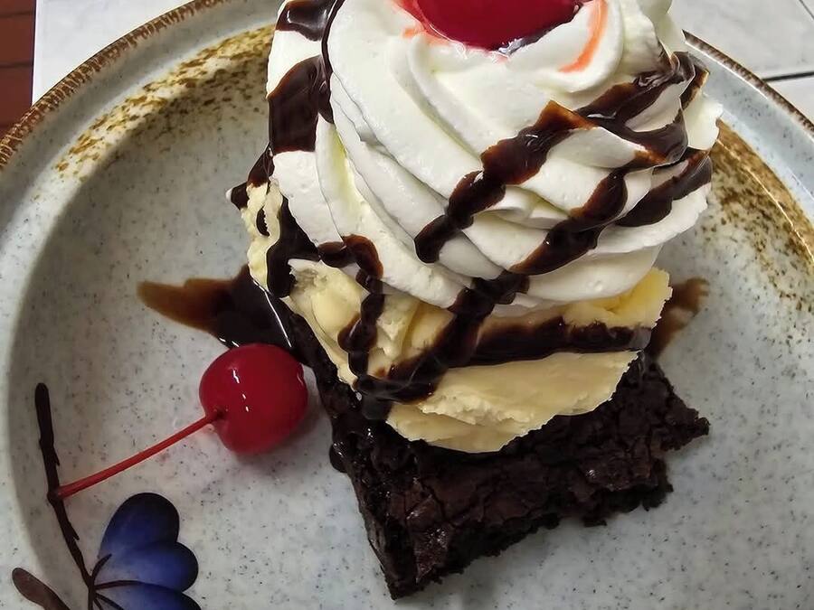 A slice of layered dessert with whipped cream, chocolate, and a cherry on top, served on a decorative plate.