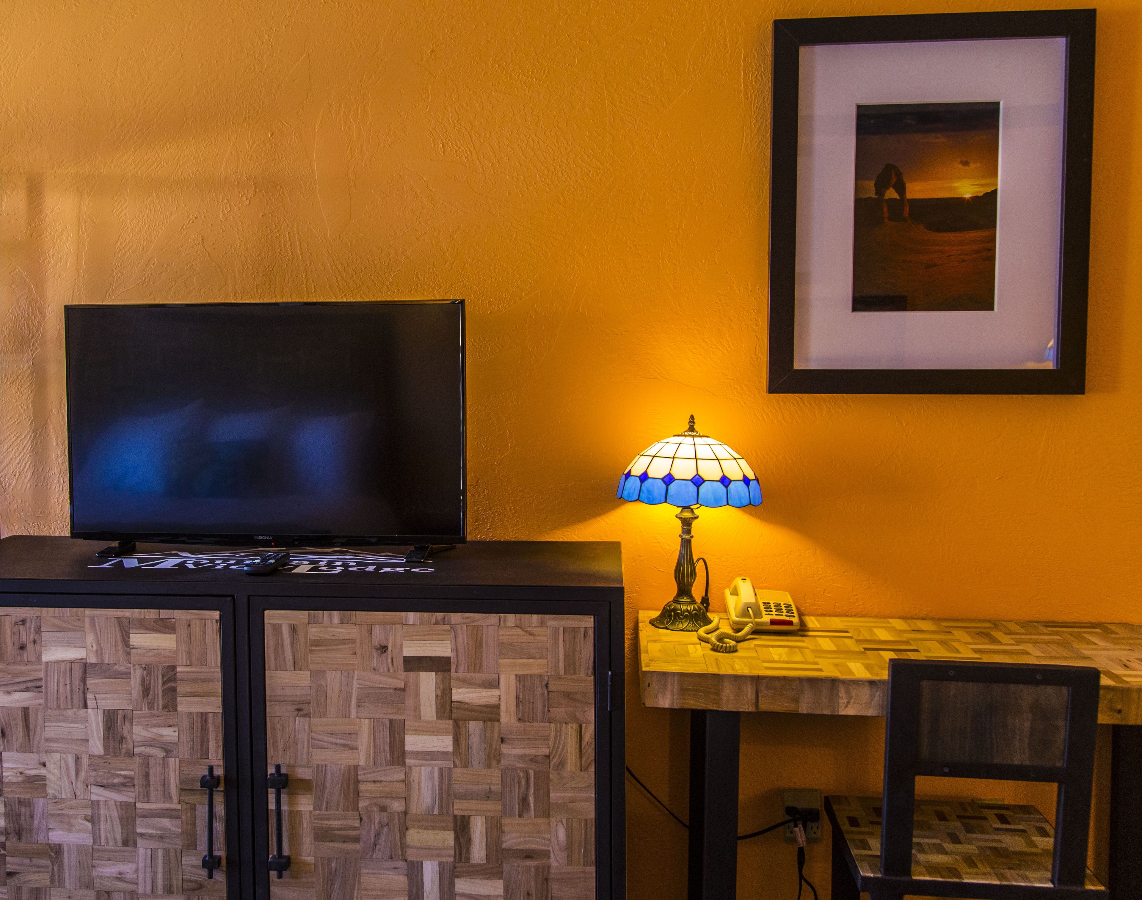 A room with a television on a cabinet, a table with a lamp, a phone, and a framed picture on the wall.