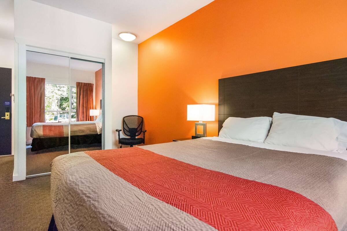 A hotel room with a large bed, a nightstand with a lamp, a chair, and a sliding door leading to another room.