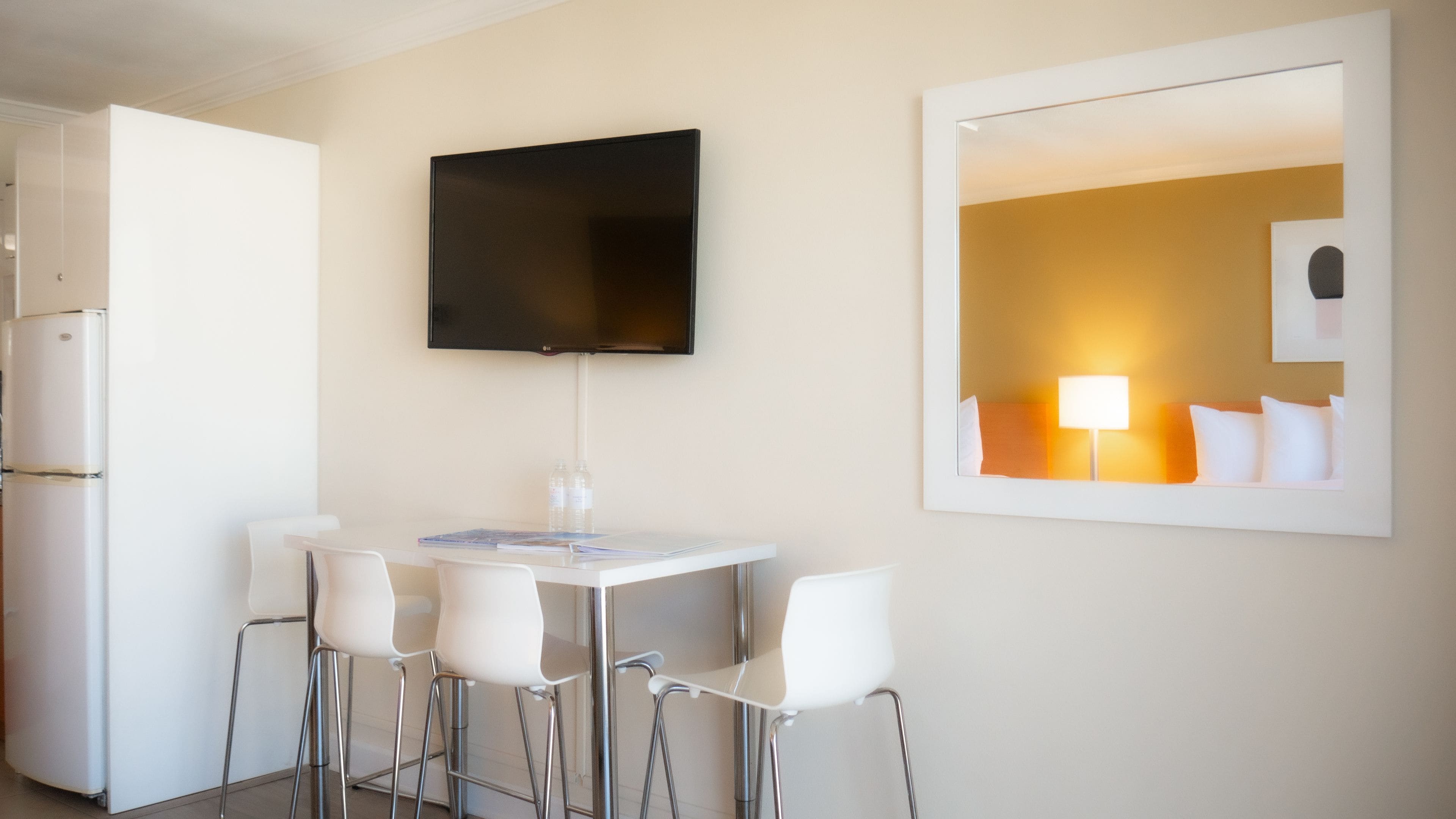 Una habitación de hotel moderna con un televisor de pantalla plana montado en la pared, una pequeña mesa de comedor con cuatro sillas blancas y un espejo grande que refleja el interior de la habitación.