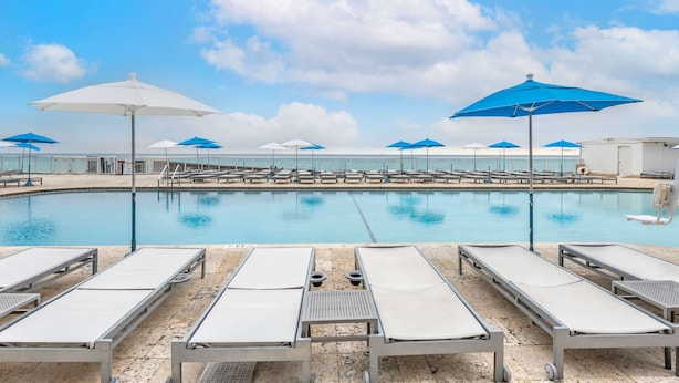 Un área junto a la piscina con sillas de descanso y sombrillas, cielo despejado y una piscina tranquila.