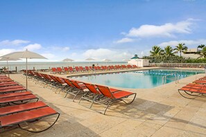 A poolside area with rows of red lounge chairs and umbrellas, a clear blue sky, and a calm sea in the background. - Ramada Plaza by Wyndham Marco Polo Beach Resort (Sunny Isles Beach)