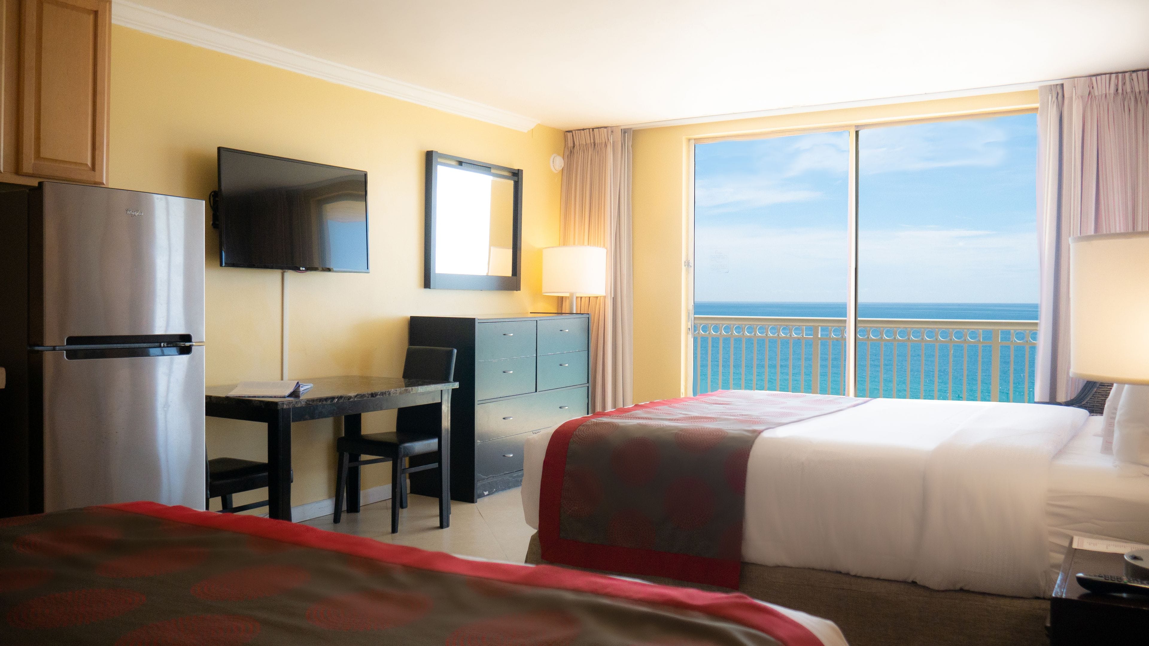 Habitación de hotel con cama, escritorio, televisión, refrigerador y balcón con vista al mar.