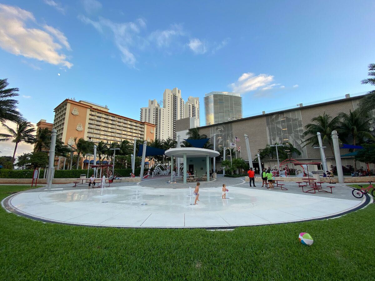 A water park with a central fountain, surrounded by palm trees and a building in the background.
