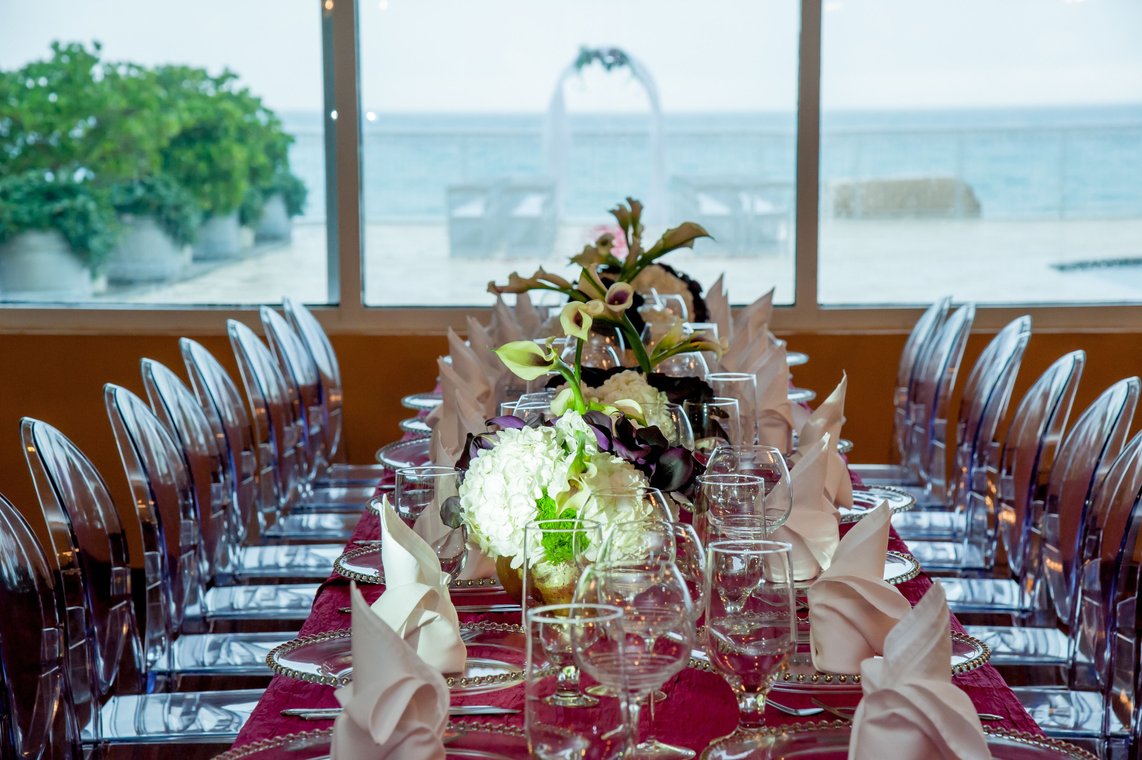 Una mesa dispuesta para un evento formal con sillas de cristal transparente, mantel rojo y un centro de mesa floral.