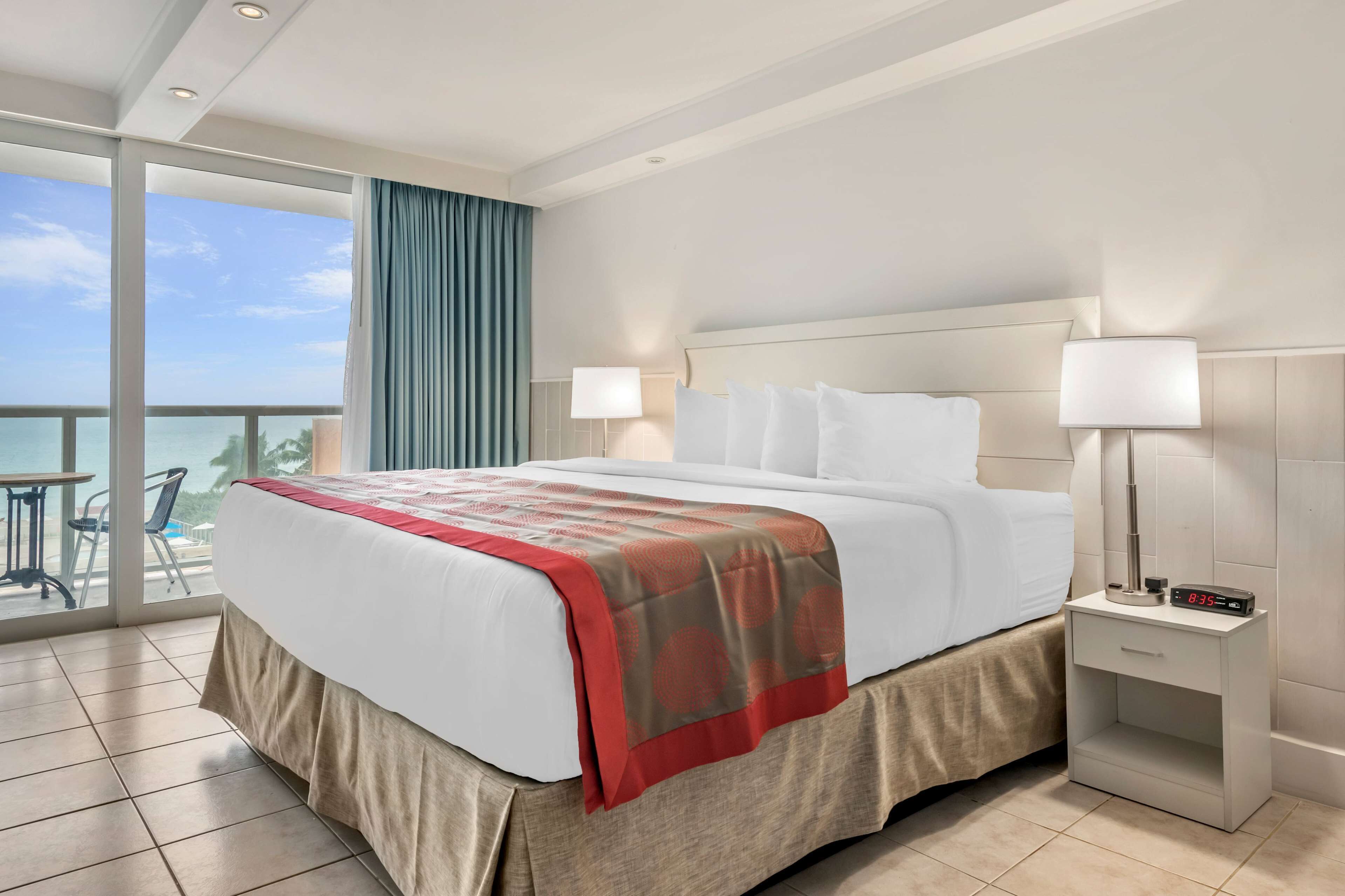 A hotel room with a large bed, two bedside tables, a view of the ocean, and a balcony with a table and chairs.