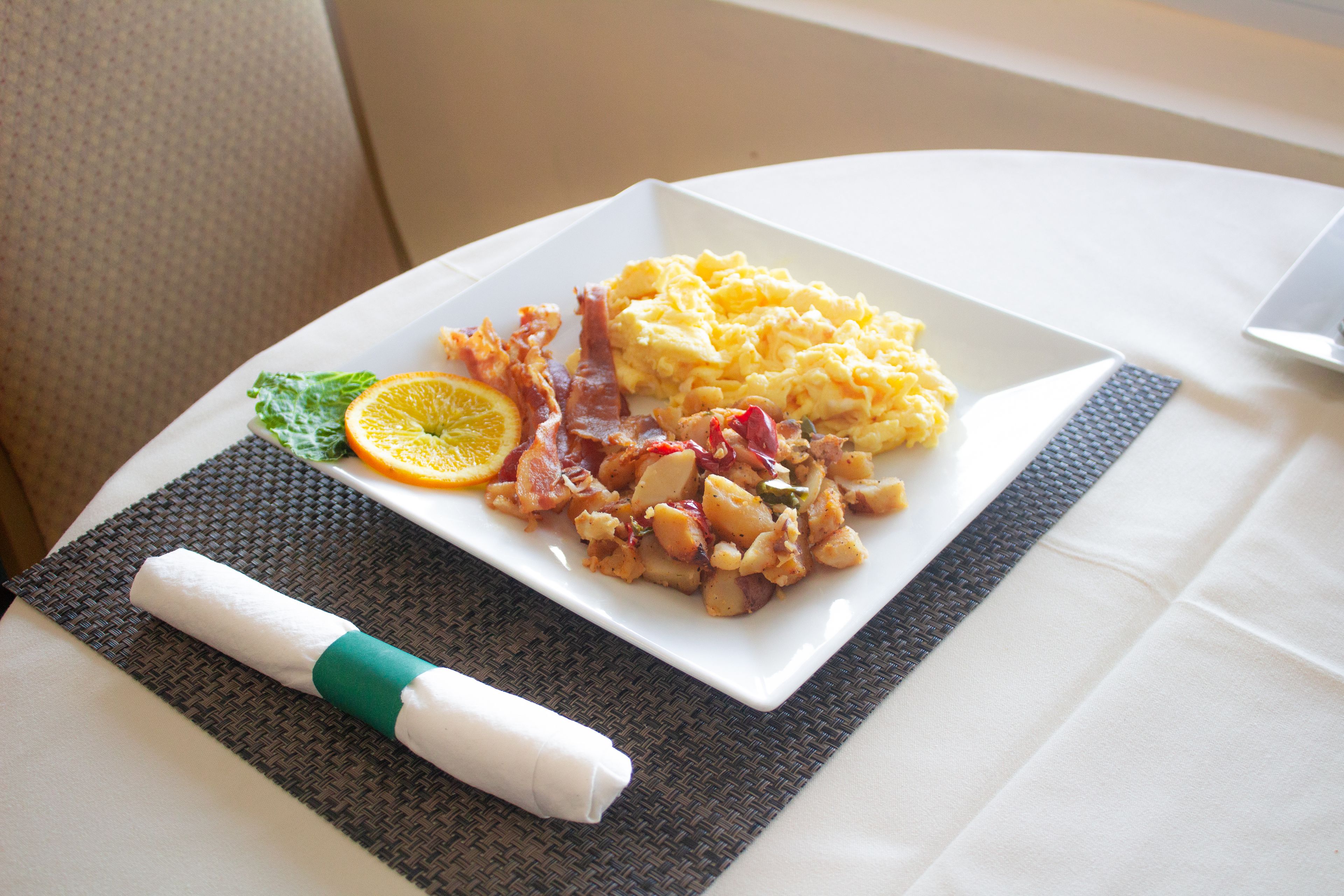 A breakfast plate with scrambled eggs, bacon, and potatoes, garnished with an orange slice and a leaf, on a white plate with a napkin and a woven placemat.