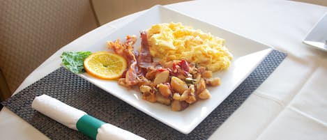 A breakfast plate with scrambled eggs, bacon, and potatoes, garnished with an orange slice and a leaf, on a white plate with a napkin and a woven placemat.