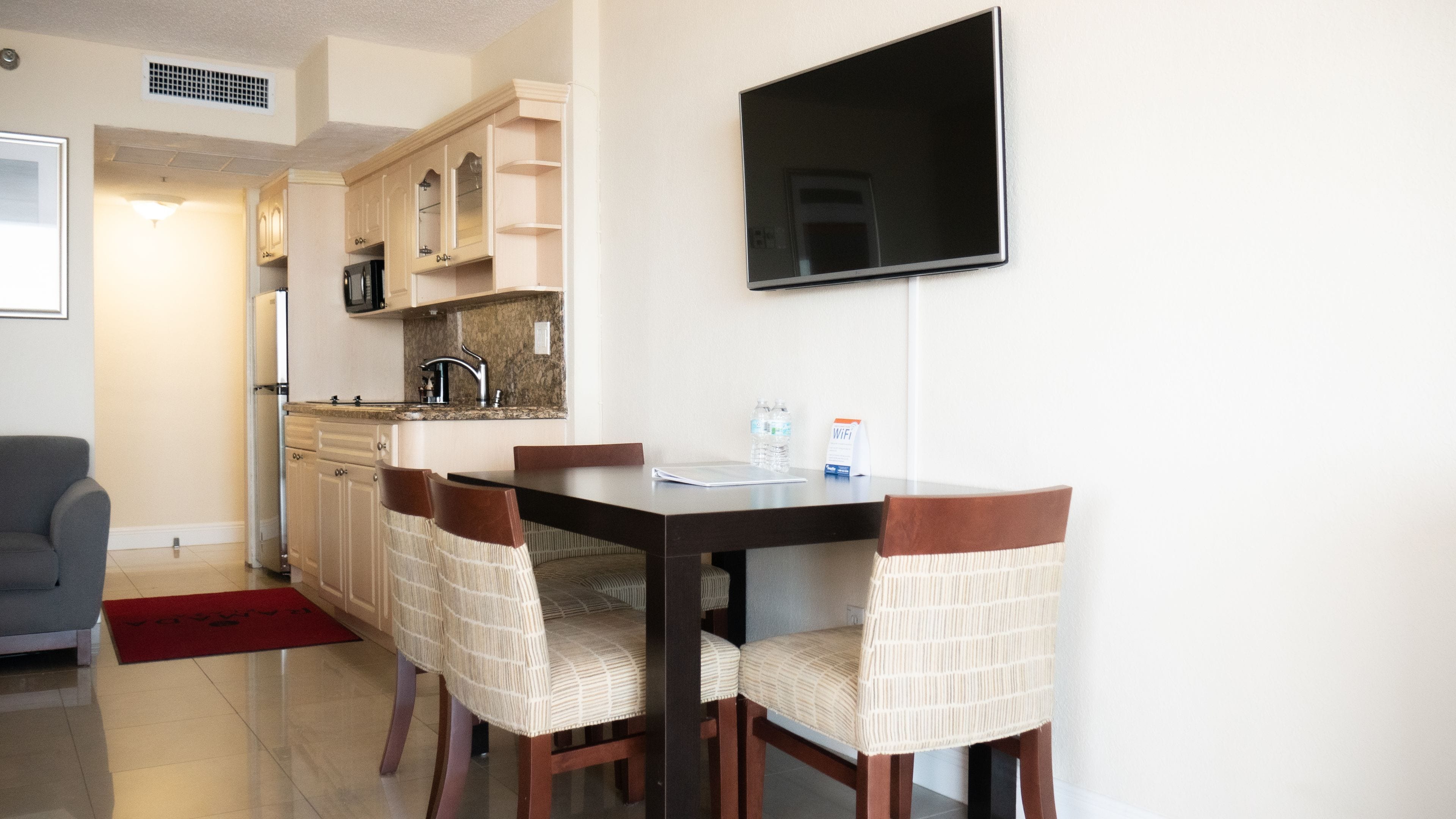 Un comedor con una mesa y cuatro sillas, un televisor de pantalla plana montado en la pared y una cocina al fondo.