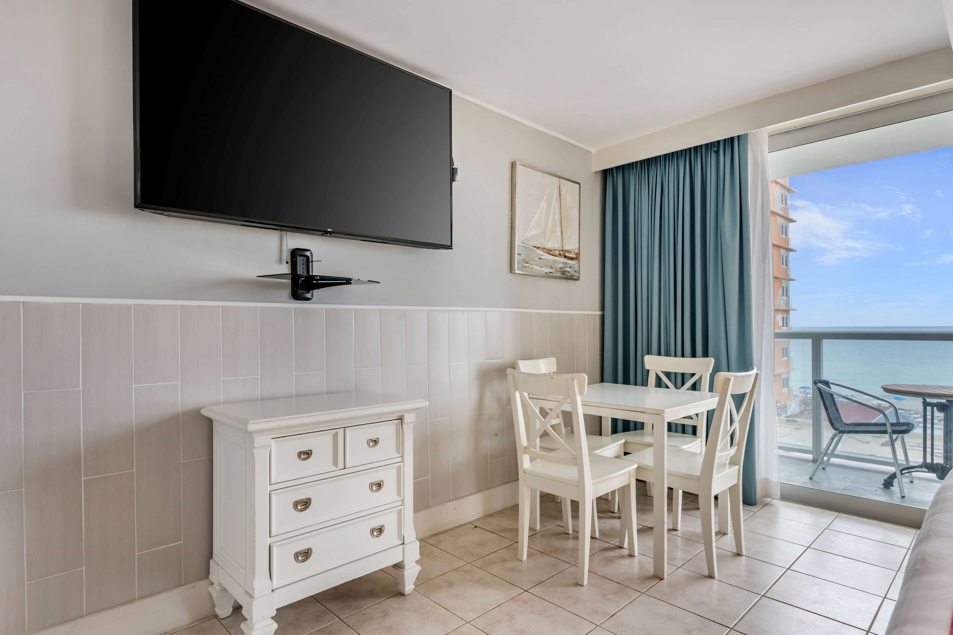 A hotel room with a flat-screen TV mounted on the wall, a white dresser with drawers, a dining table with chairs, and a balcony with a view of the sea and buildings.