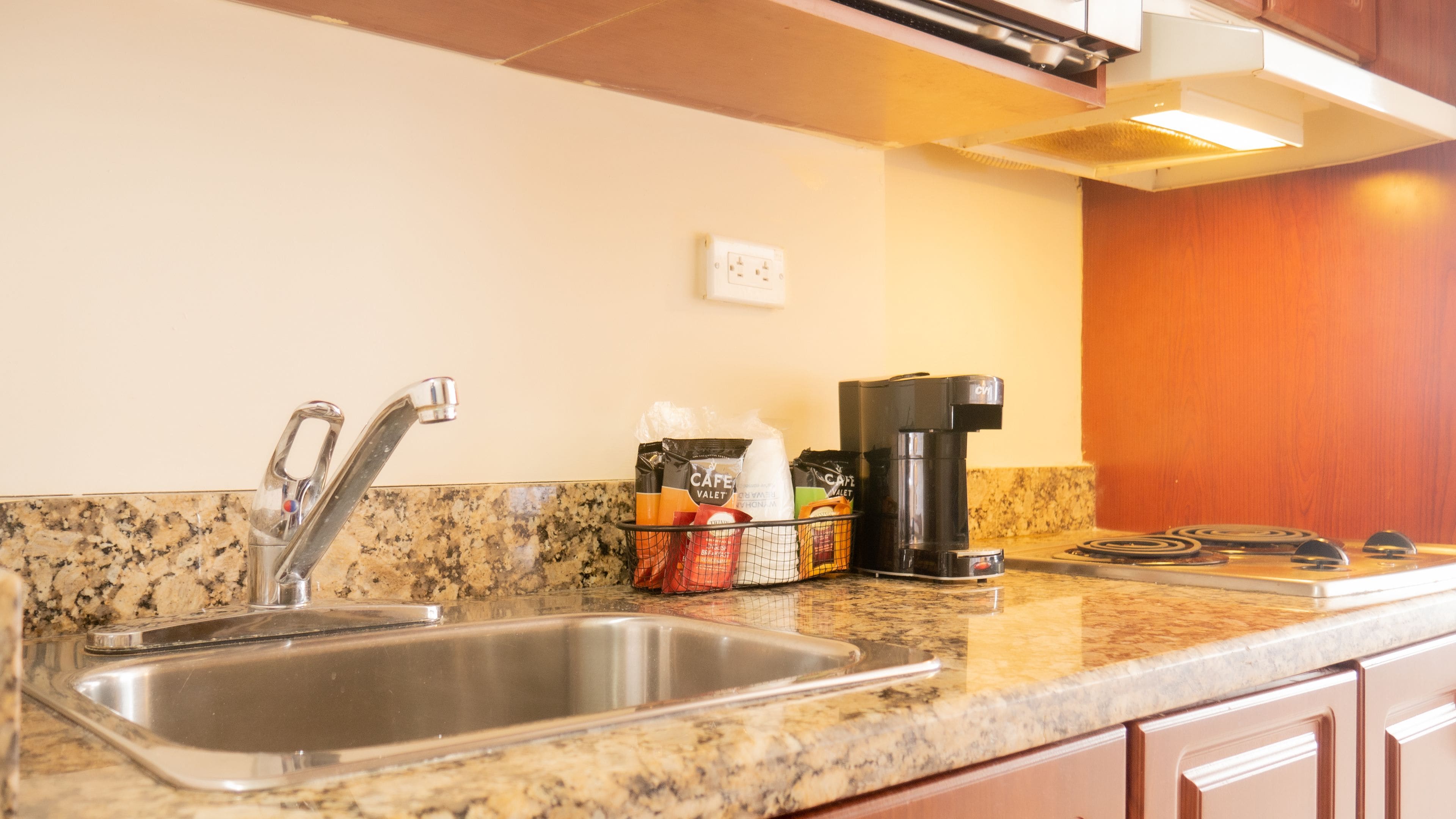 A kitchen with a granite countertop, stainless steel sink, coffee maker, and a basket of coffee pods.