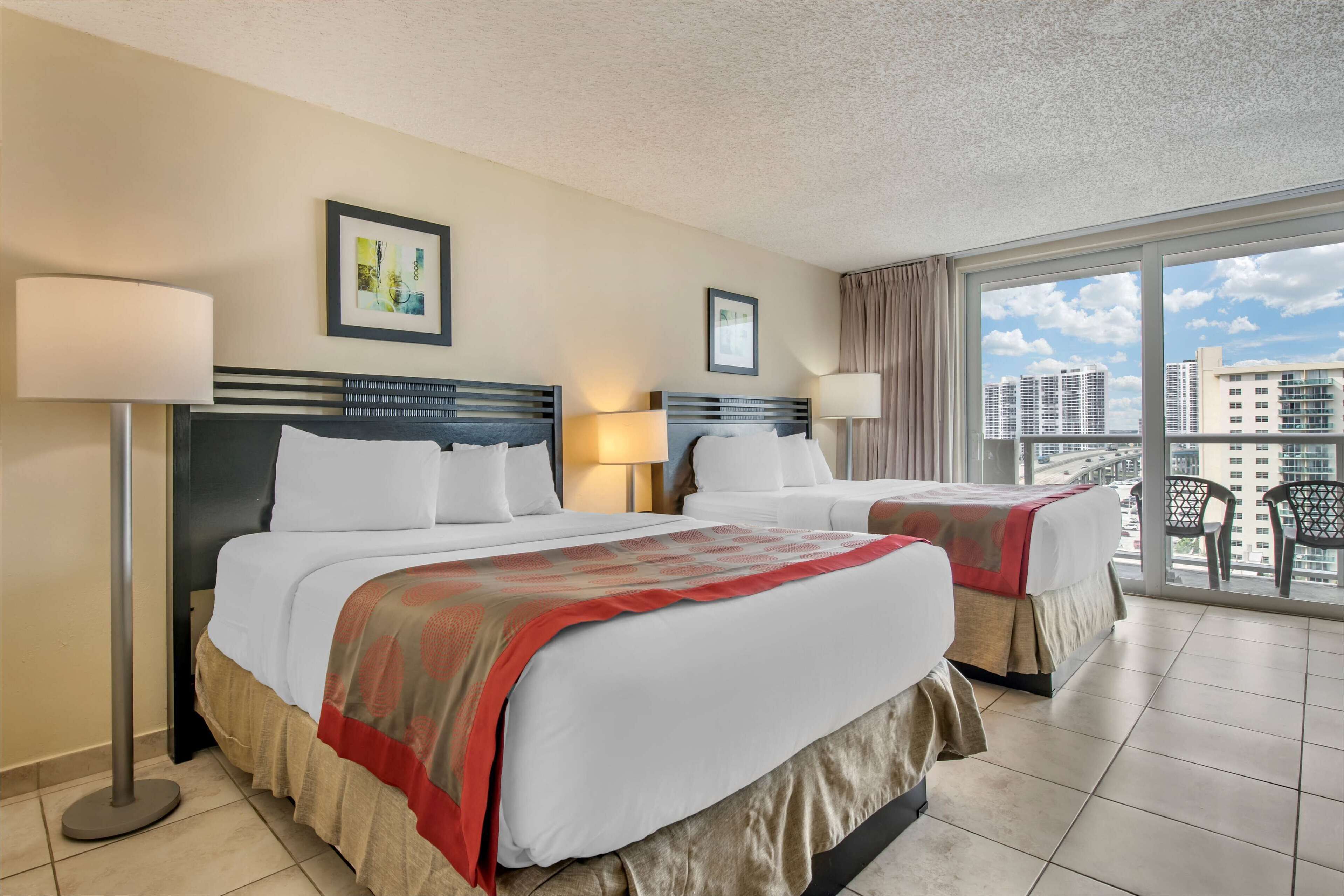 A hotel room with two beds, a large window with a city view, and a balcony with chairs.