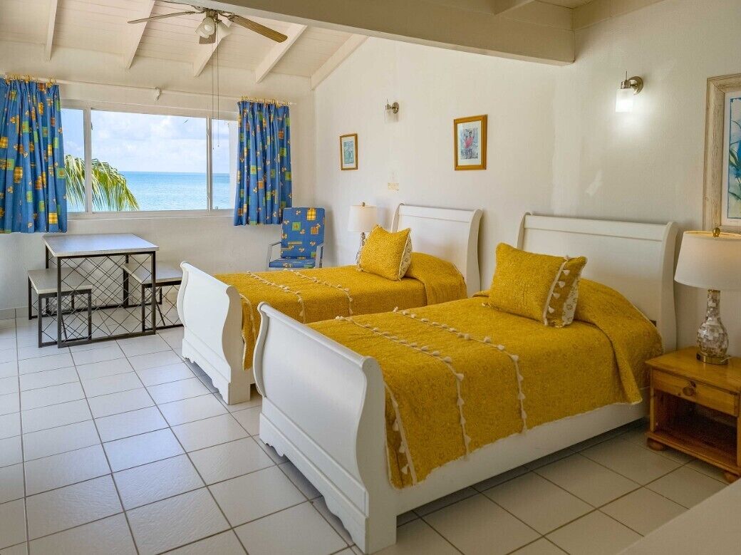 A room with two beds, a table, a chair, a lamp, and a view of the ocean.