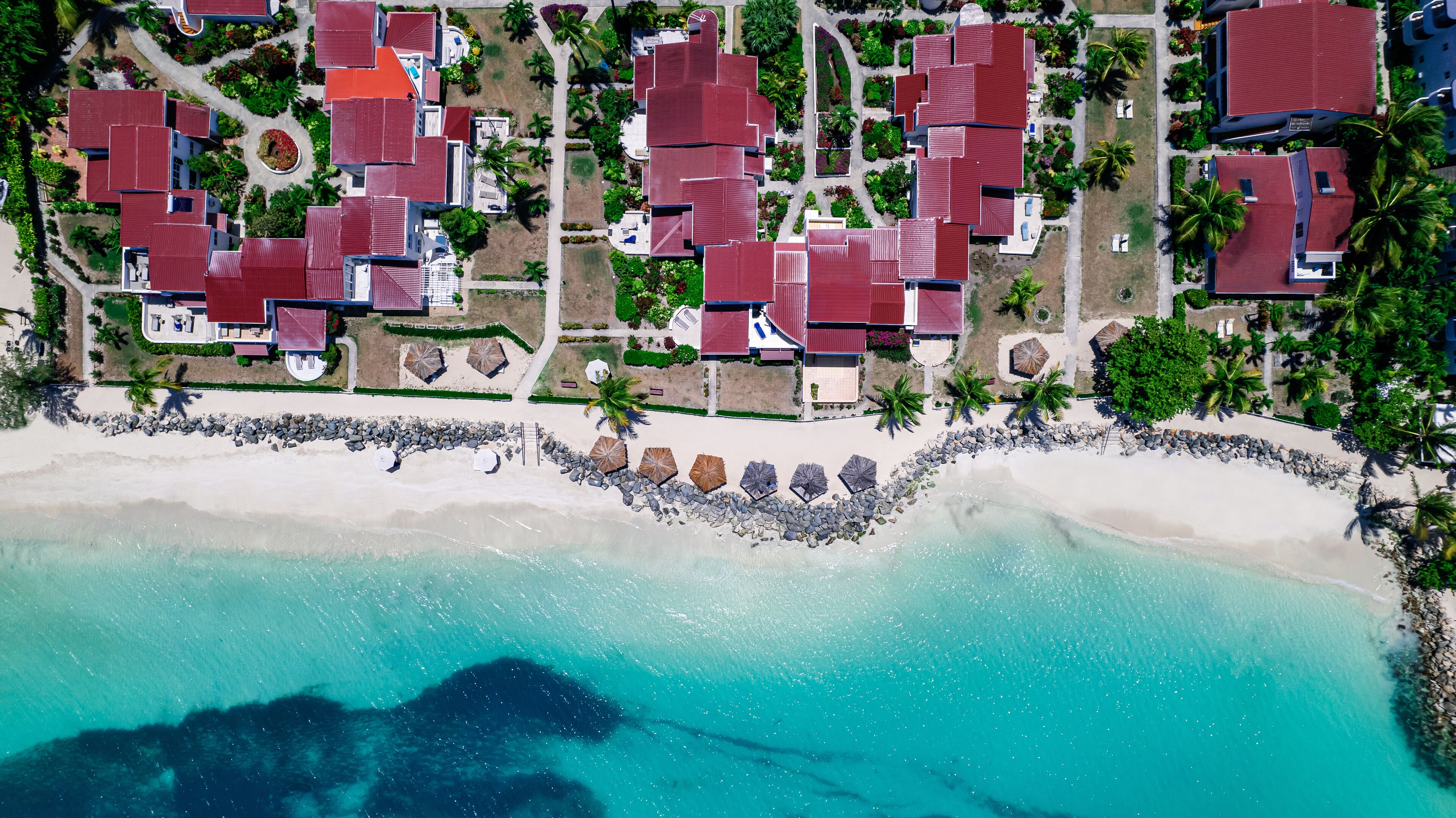 Vista aérea de un complejo turístico costero con varios edificios de tejados rojos, una playa de arena y aguas turquesas cristalinas.