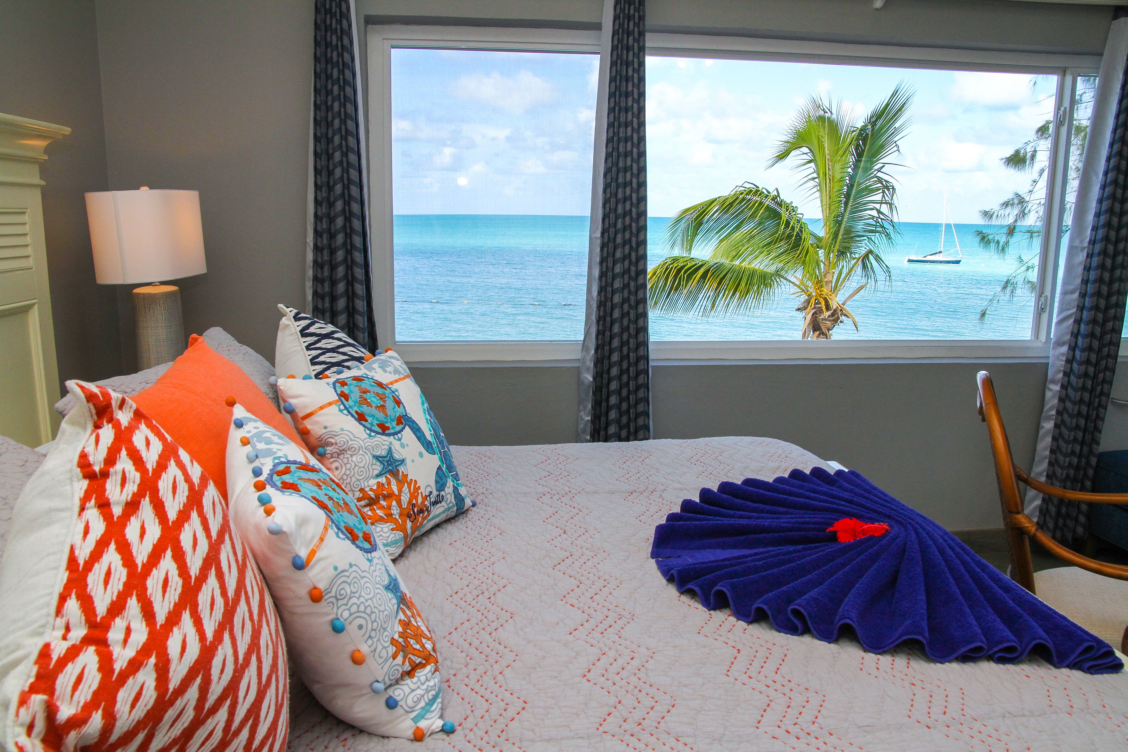 Un dormitorio con cama, almohadas, ventana y vista al océano y palmeras.