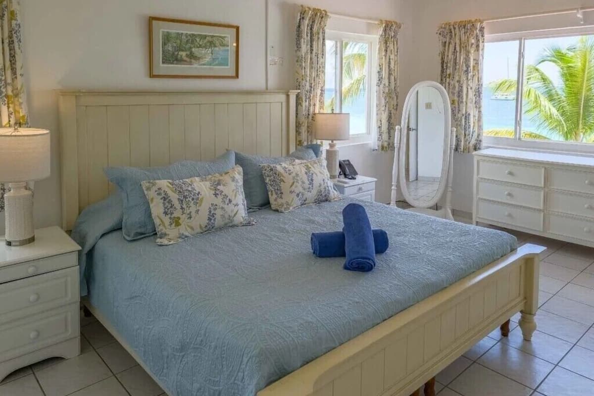 Une chambre à coucher avec un lit, une table de chevet, une commode, un miroir et une fenêtre avec des rideaux.