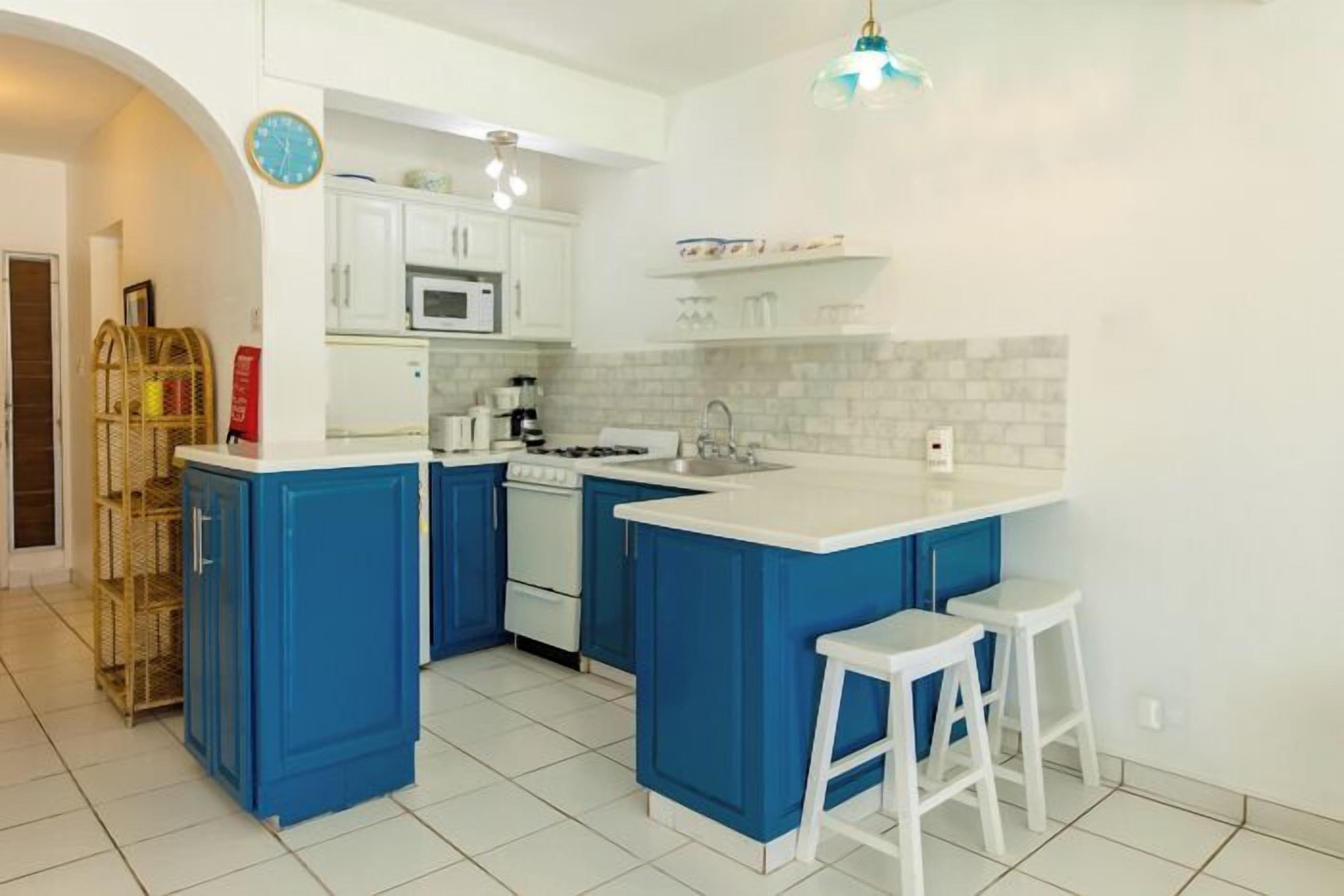 Una cocina con armarios azules, encimeras blancas, microondas, estufa y un lavamanos. Hay dos taburetes blancos y una lámpara colgante.