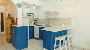 Una cocina con armarios azules, encimeras blancas, microondas, estufa y un lavamanos. Hay dos taburetes blancos y una lámpara colgante.