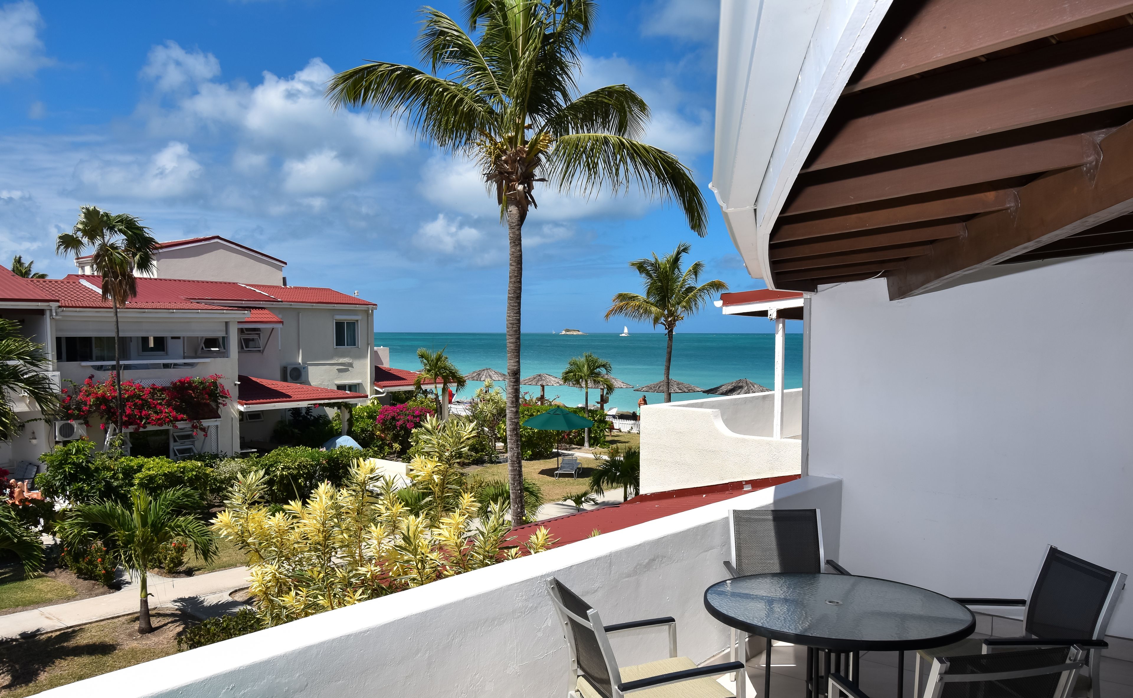 A balcony with a glass table and chairs, overlooking a beach with palm trees and a building with a red roof.