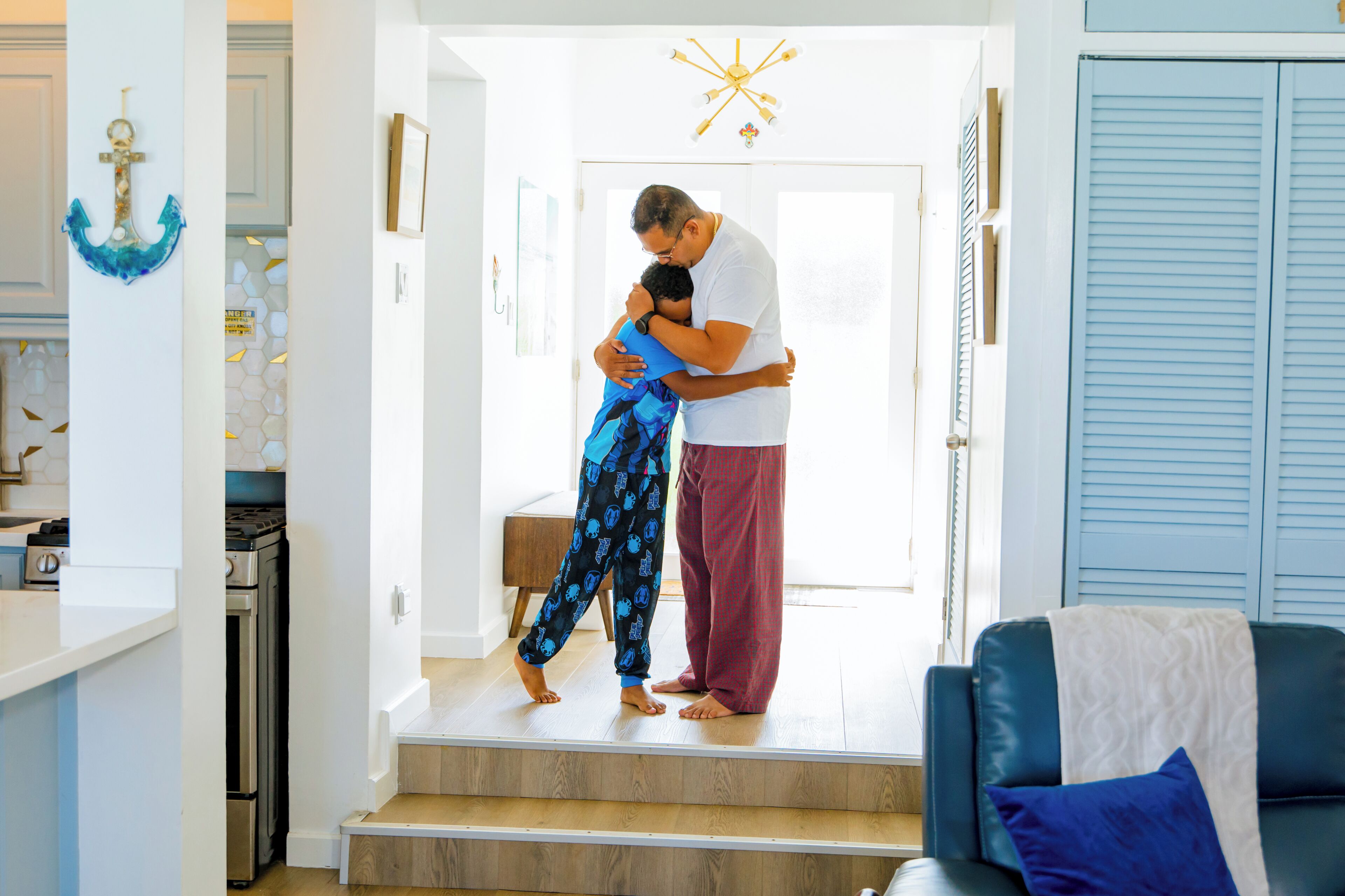 Un hombre y un niño se abrazan en un pasillo luminoso con una escalera, una zona de cocina y un sofá azul.