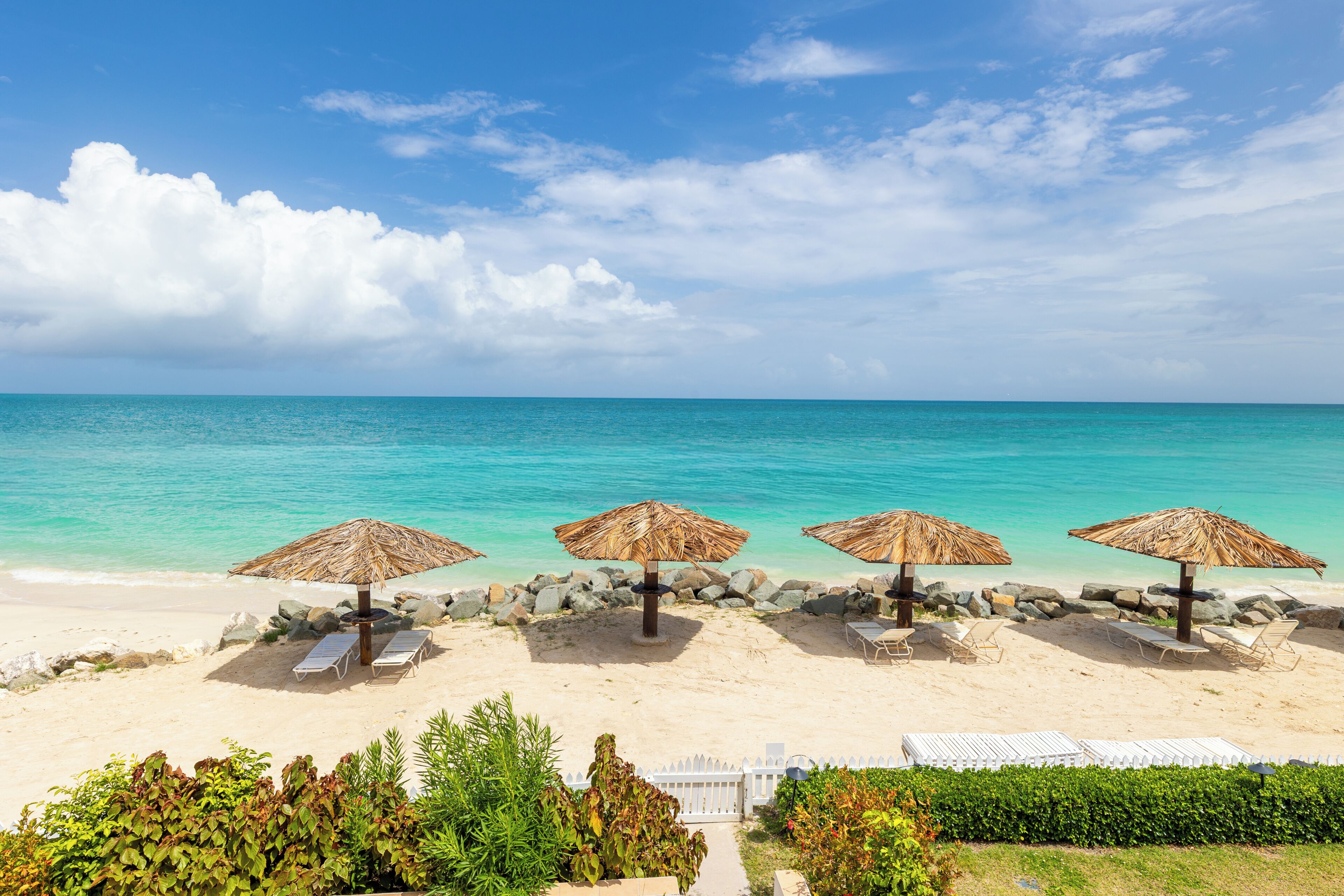 A beach with four thatched umbrellas, lounge chairs, and a white fence.