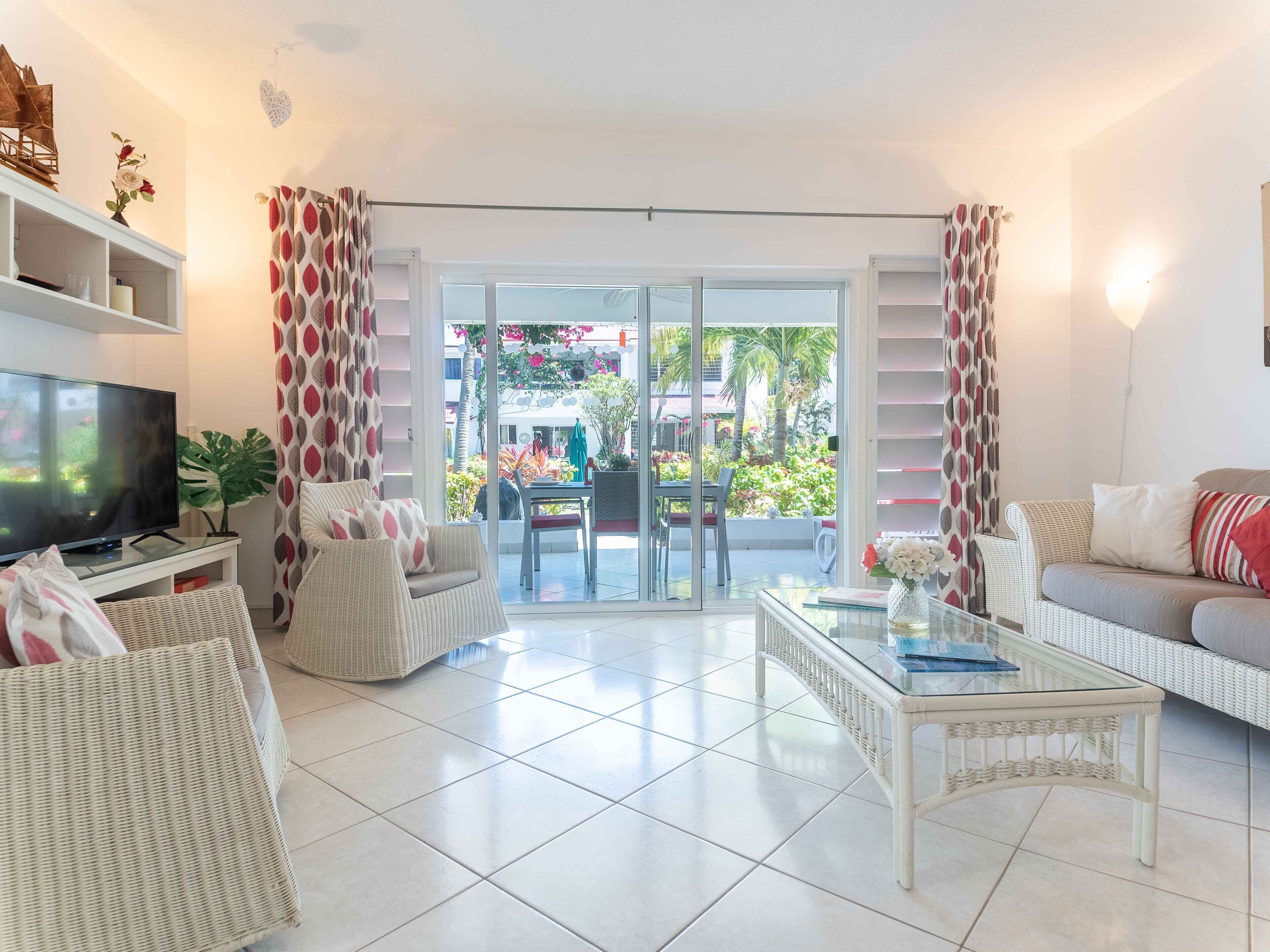 A living room with a TV, wicker furniture, a glass coffee table, and a view of a garden through glass doors.