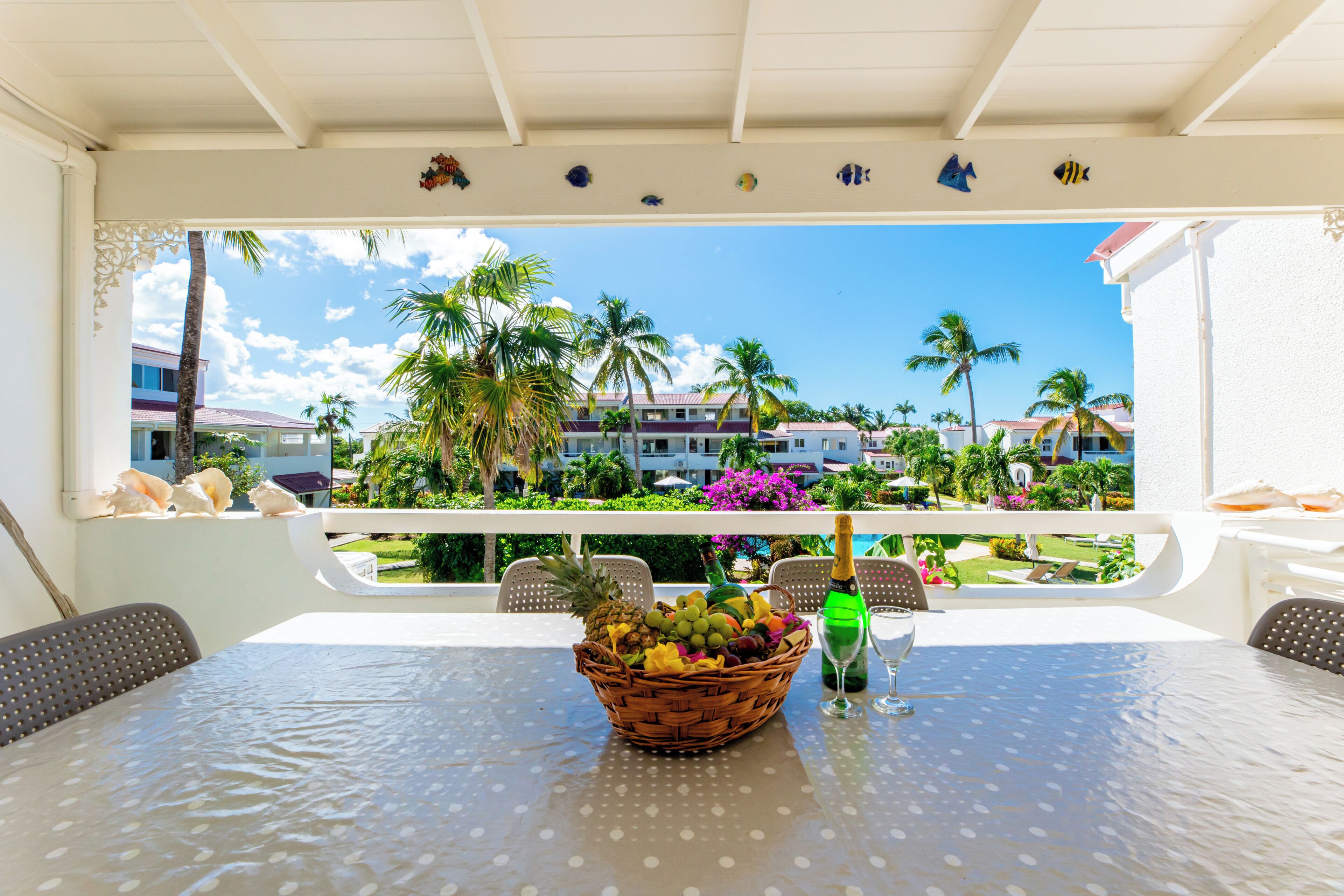 A table with a fruit basket and champagne, overlooking a pool and palm trees.