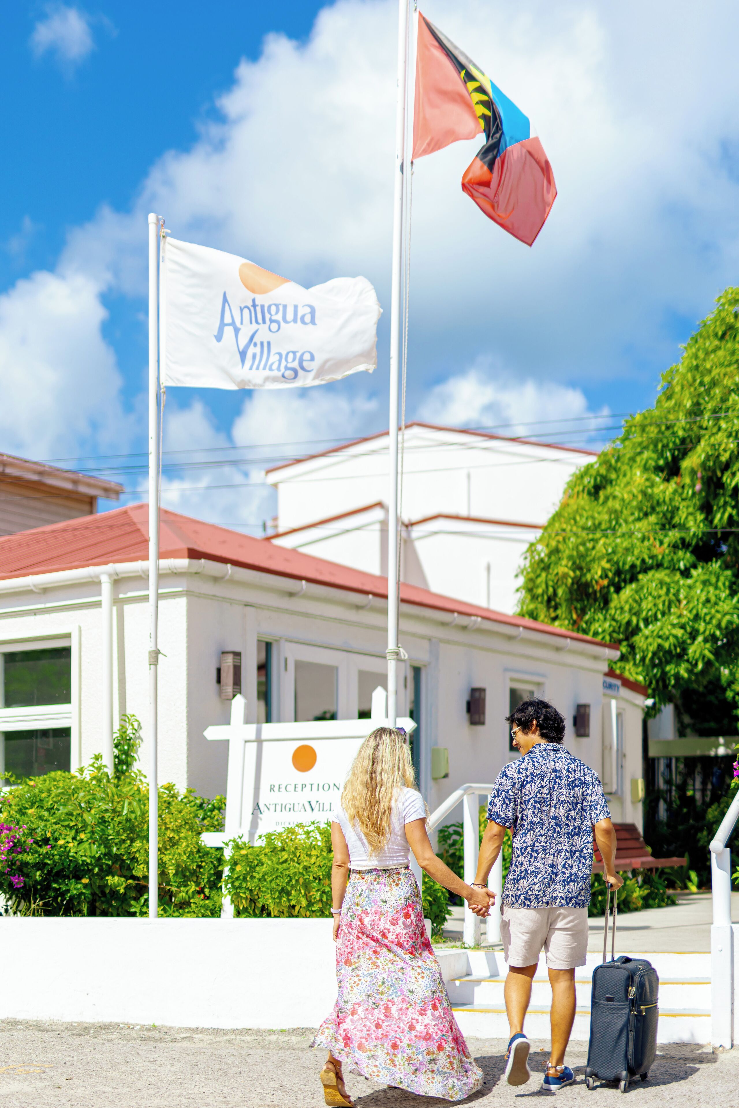A couple holding hands in front of a building with a flag labeled "Antigua Village."