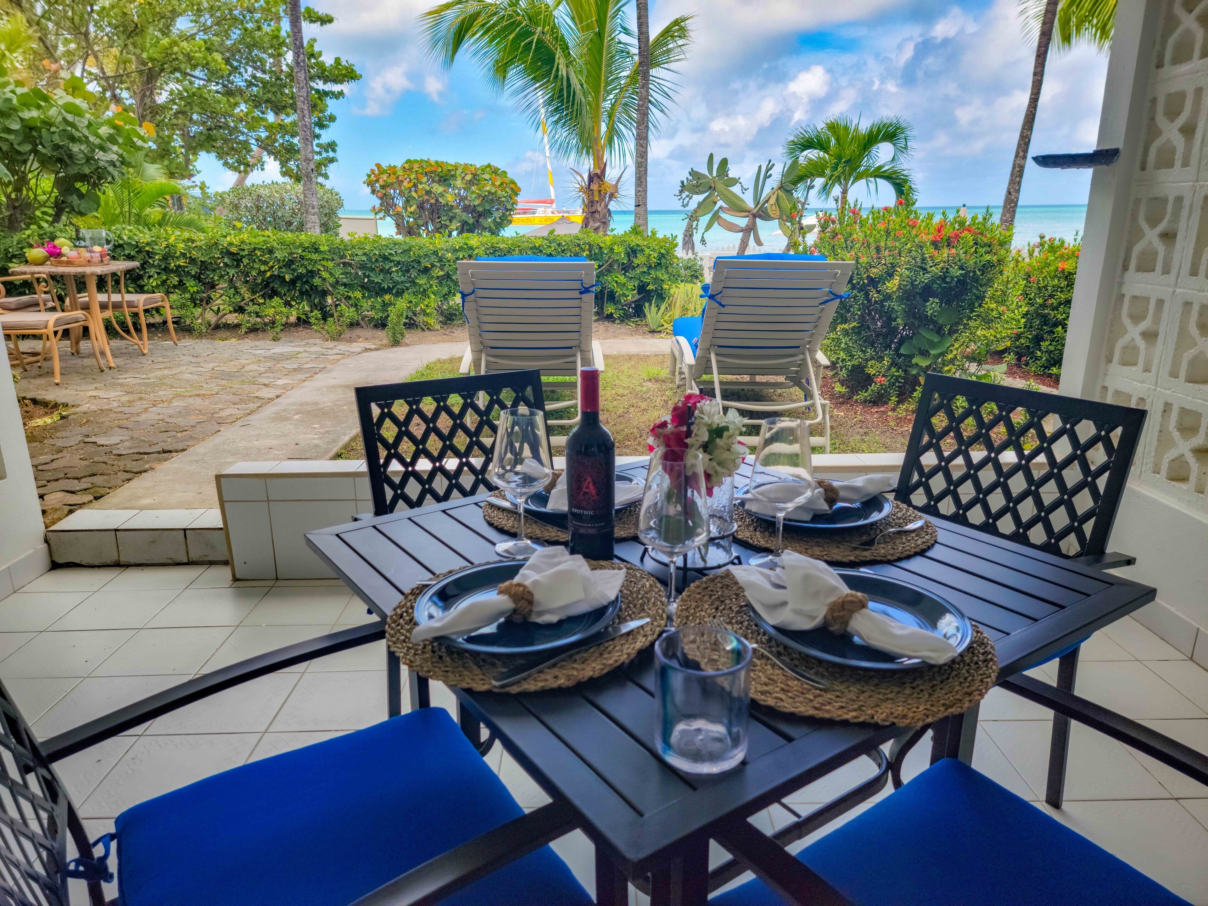 Un espace de restauration en plein air avec une table dressée pour deux personnes, entourée de chaises, et offrant une vue sur une plage bordée de palmiers et un ciel dégagé.