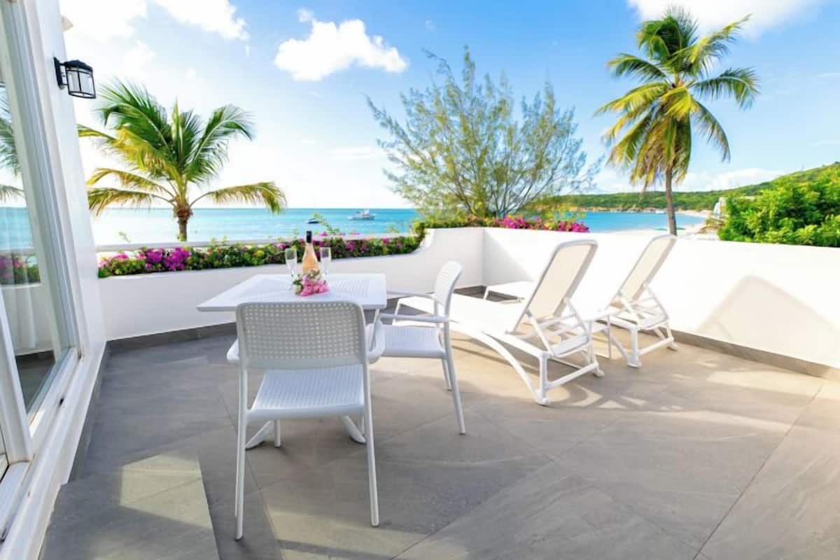 Un balcon avec une table et des chaises, donnant sur une plage bordée de palmiers et offrant une vue sur un ciel dégagé.