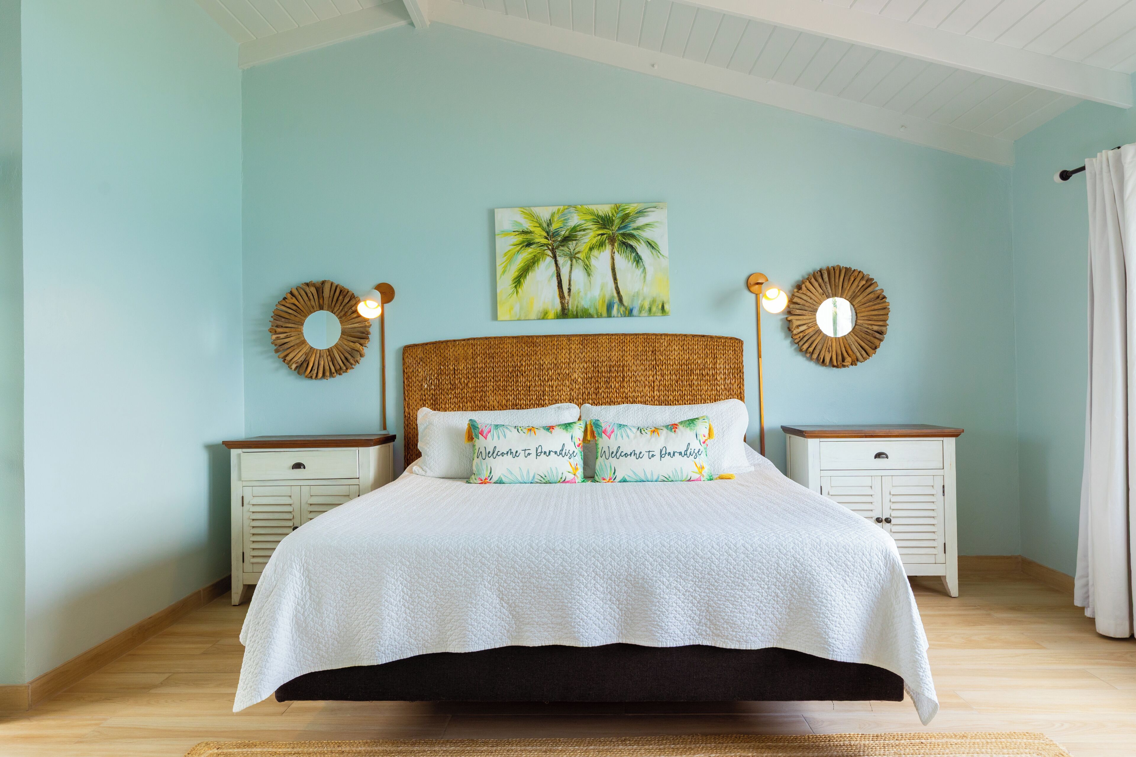 A bedroom with a bed, two nightstands, a painting of palm trees, and two wall sconces.