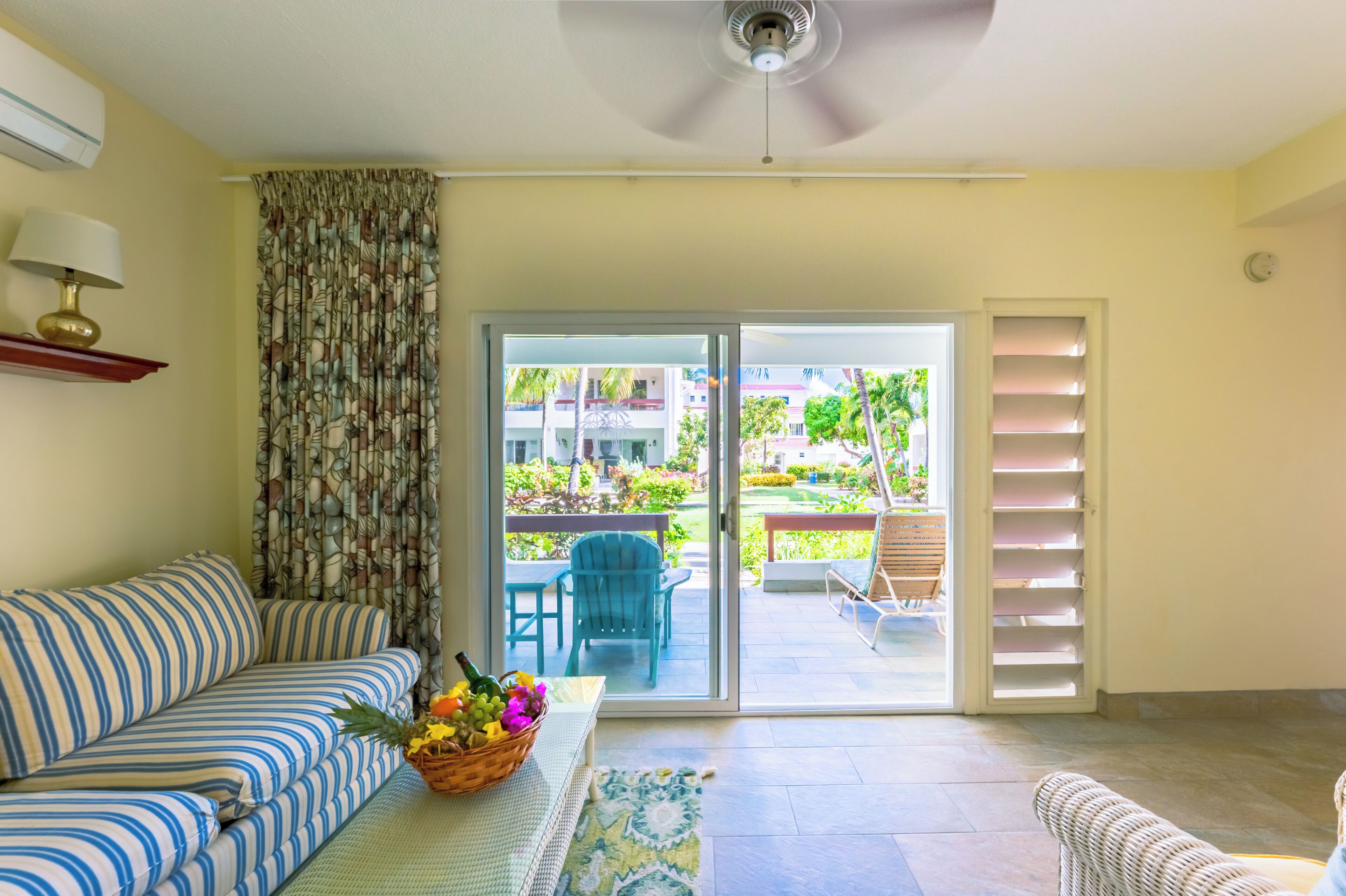 A living room with a sofa, a table with a fruit basket, a ceiling fan, and a sliding glass door leading to an outdoor area with a chair and a table.