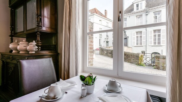 A dining table set with white tableware, a salt and pepper shaker, and a potted plant. A window reveals a view of a white building and a cobblestone street. - Bryghia Hotel (Bruges)