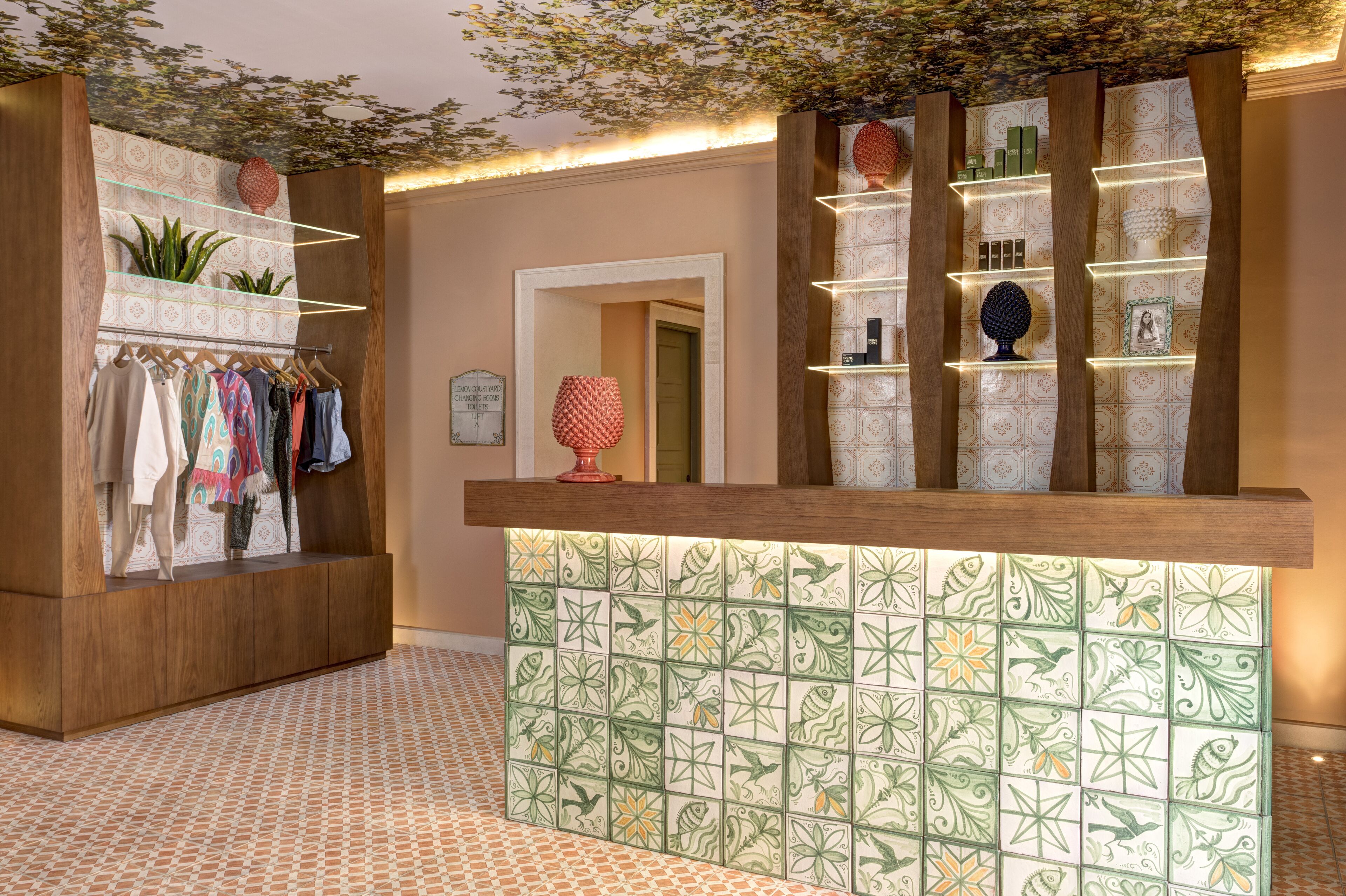 Uma área de recepção de hotel com balcão de madeira, parede revestida com padrão de peixes e um cabideiro com diversas peças de roupa.