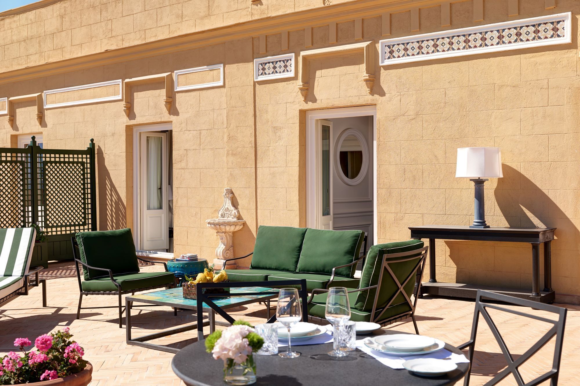 An outdoor seating area with green cushioned chairs, a round table set with plates and glasses, and a decorative fountain.