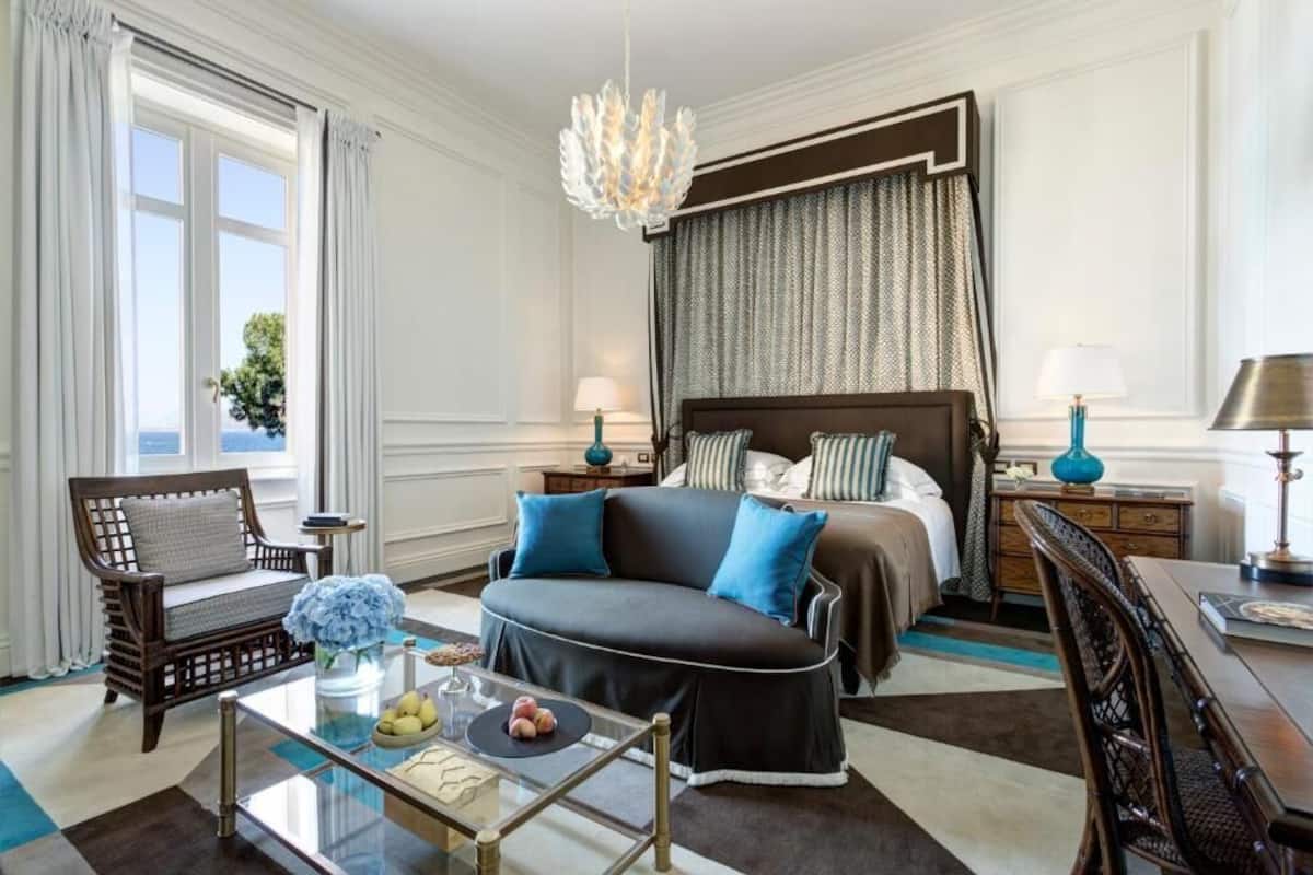 Habitación de hotel con una cama grande, una zona de estar con sofá y sillones, una mesa de centro de vidrio y vistas al exterior.