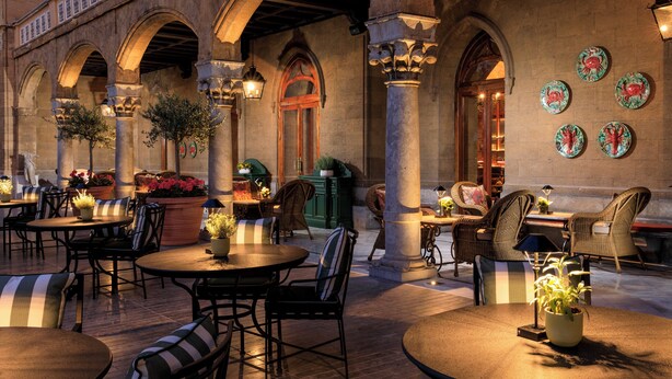 An outdoor seating area with round tables and chairs, surrounded by stone arches and columns.