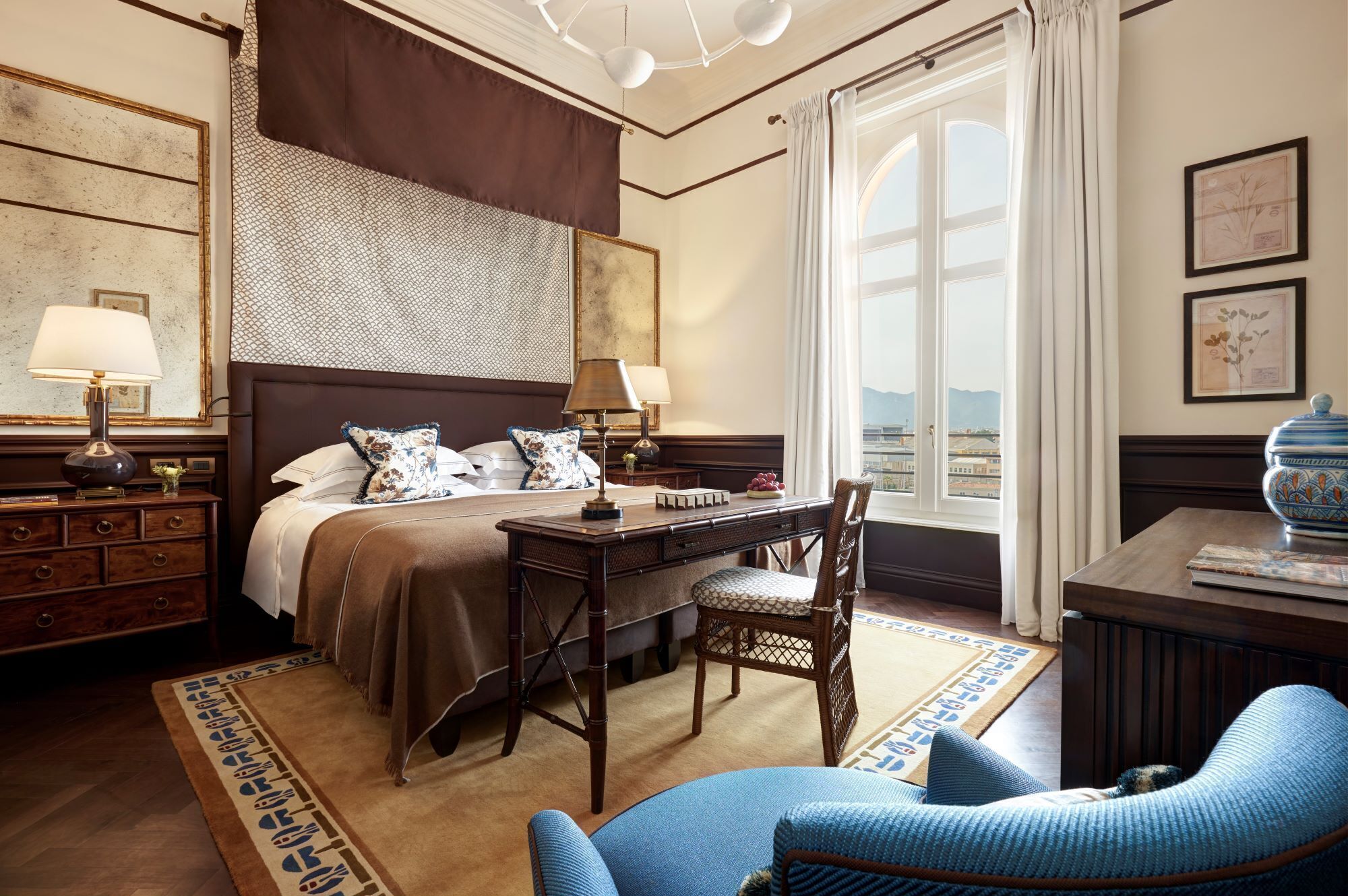 Una habitación de hotel con una cama grande, dos lámparas de noche, un escritorio con una silla, una ventana con vistas y un jarrón decorativo sobre una mesita.
