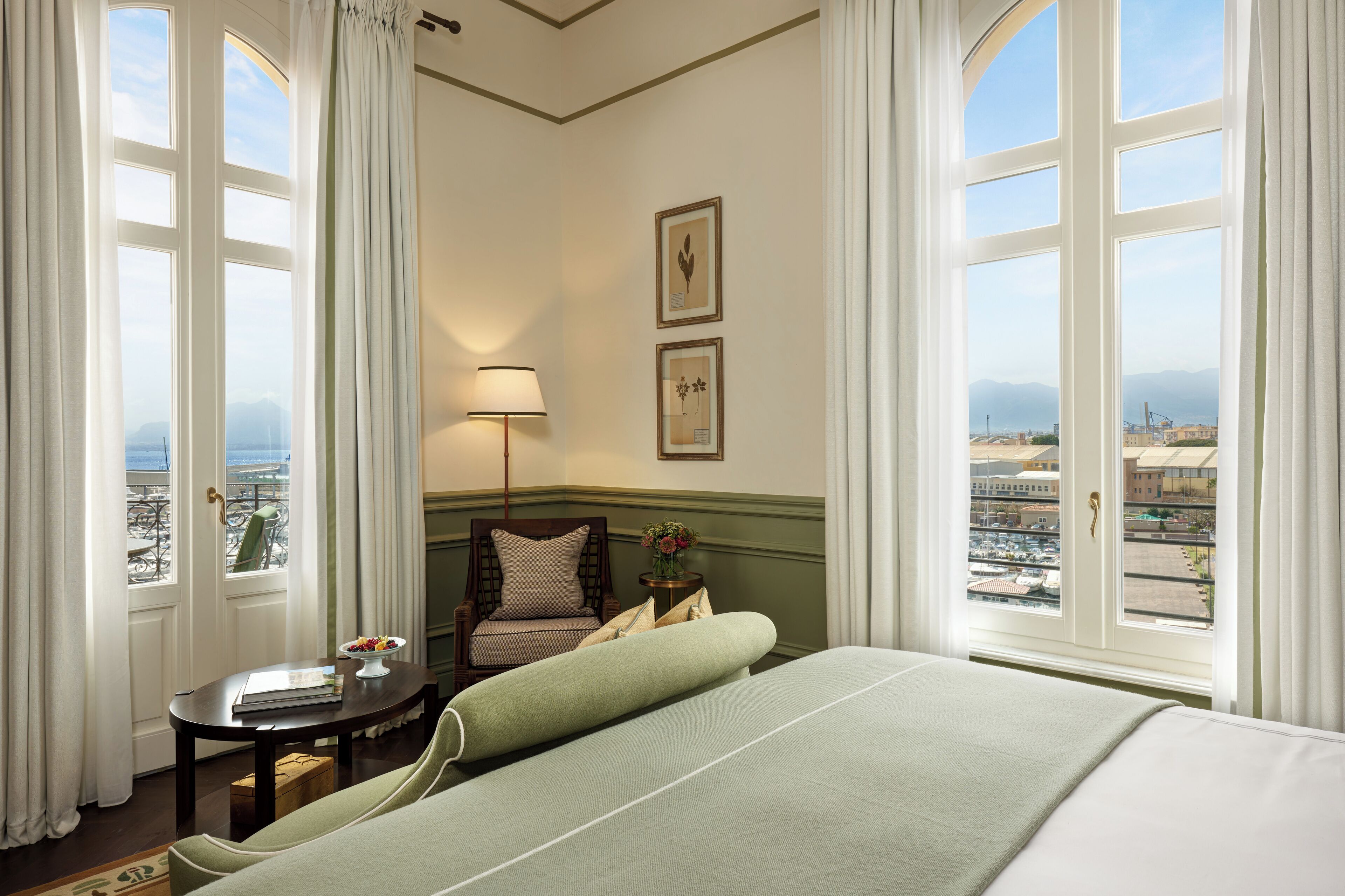 A hotel room with a bed, a chair, a small table with a lamp, and a view of the city through large windows.