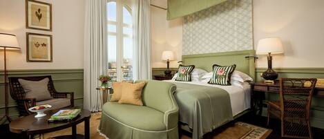 A hotel room with a bed, a green sofa, a wooden chair, a small table with a lamp, and a view of the city through a window.
