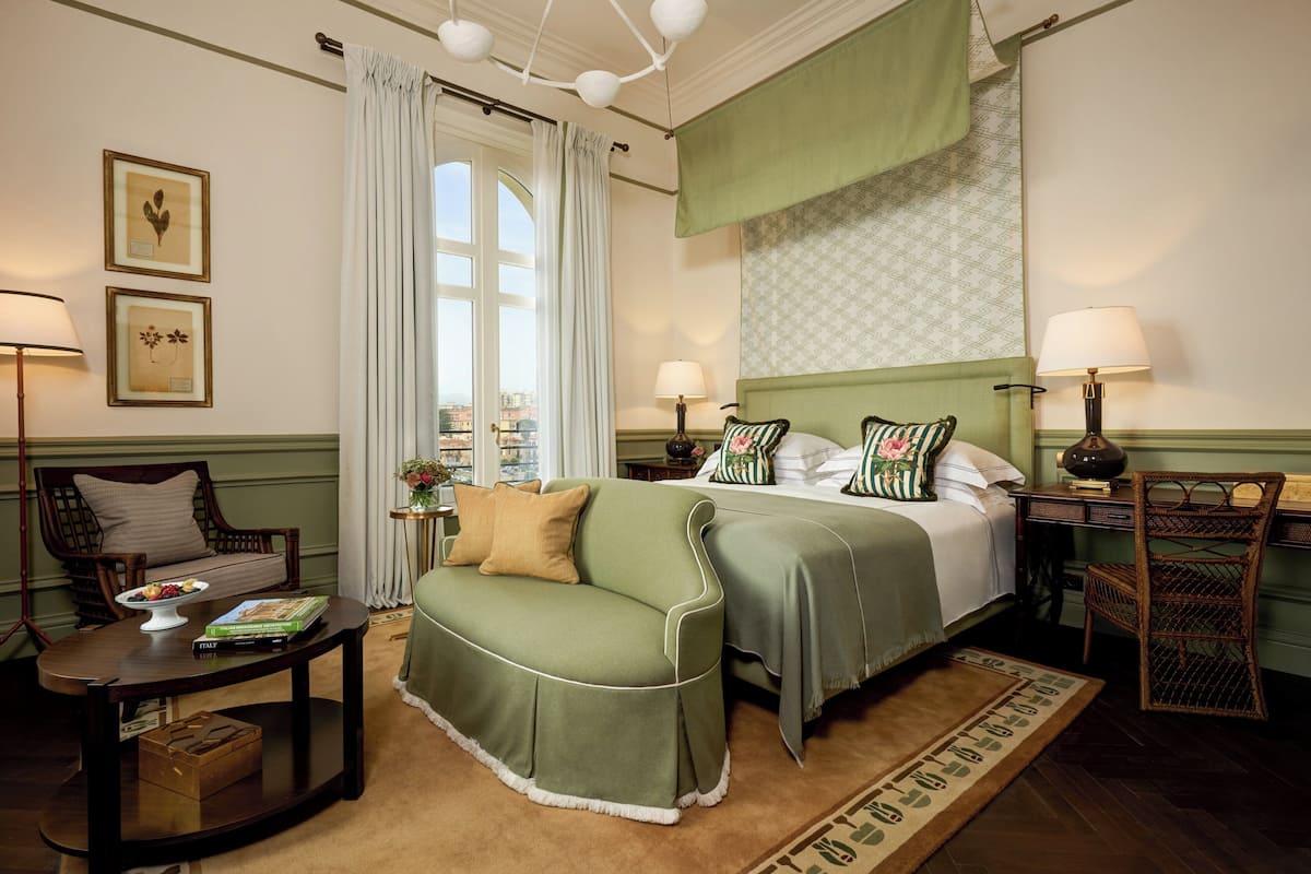 A neatly arranged hotel room with a bed, a green sofa, a wooden chair, a small table with a bowl of fruit and books, and a view of the city through a large window.