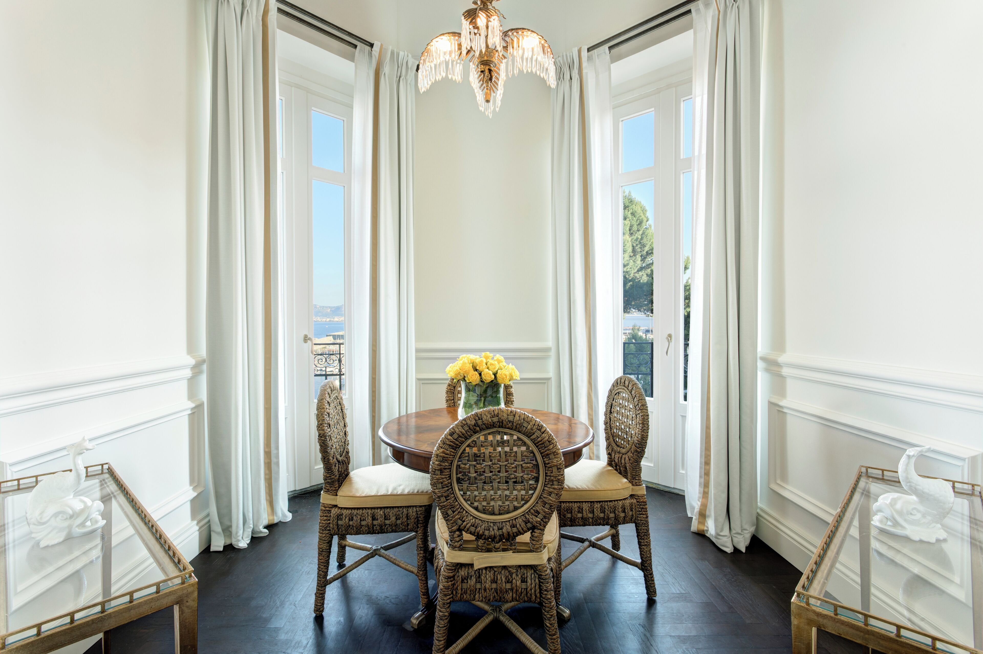 A dining area with a round table, four wicker chairs, a vase of yellow flowers, and a chandelier.