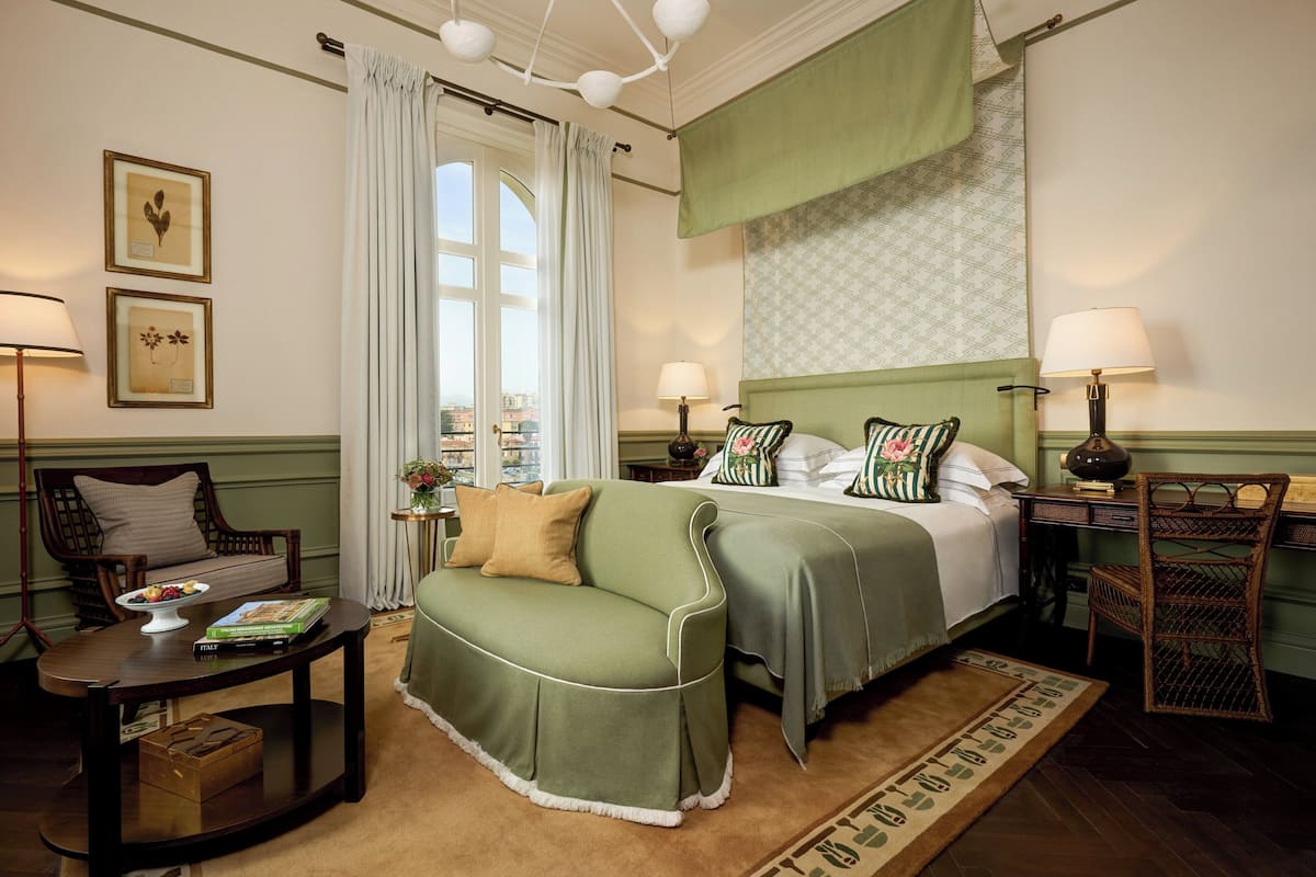 Ein ordentlich eingerichtetes Hotelzimmer mit einem Bett, einem grünen Sofa, einem Holzstuhl, einem kleinen Tisch mit einer Obstschale und Büchern sowie Blick auf die Stadt durch ein großes Fenster.