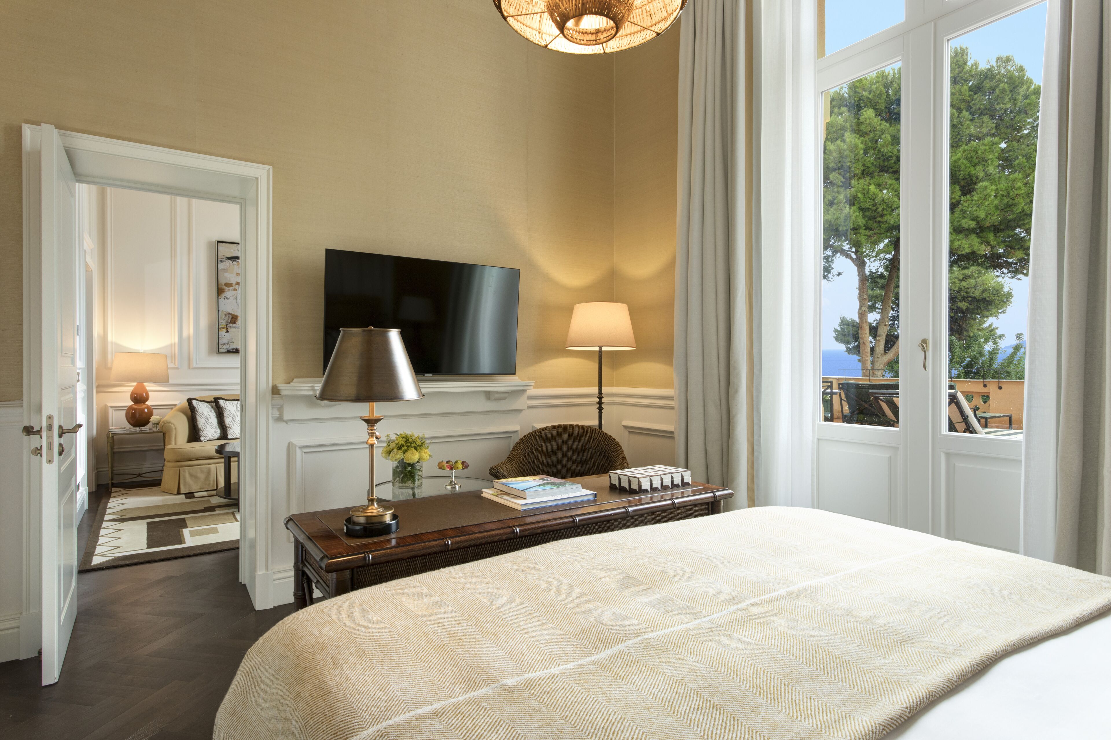 Quarto de hotel com uma cama grande, uma TV de tela plana, uma escrivaninha com abajur e vista para árvores pela janela.