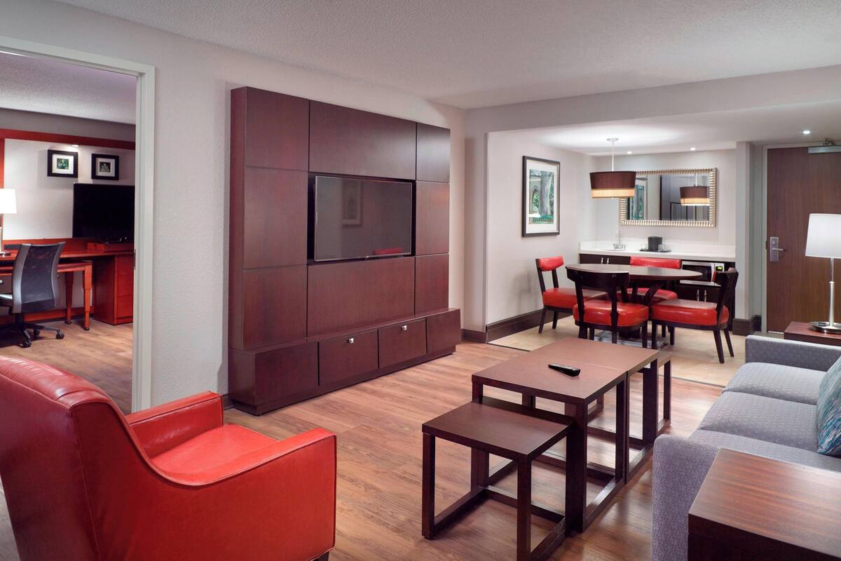 A modern hotel room with a flat-screen TV mounted on a wooden panel, a dining area with a table and chairs, and a seating area with a sofa and armchairs.